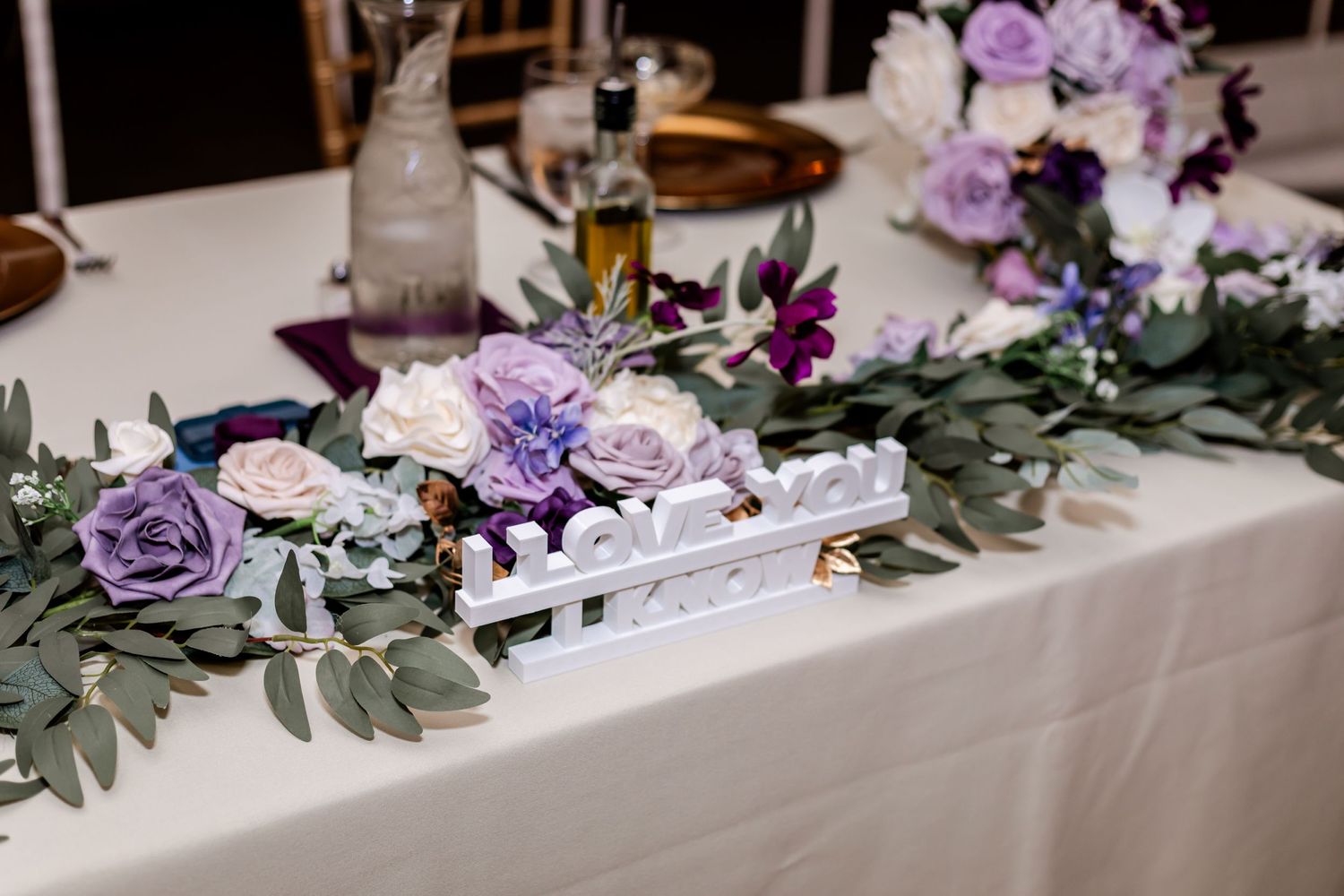 Wedding table centerpiece with purple and pink flowers and eucalyptus on white tablecloth.
