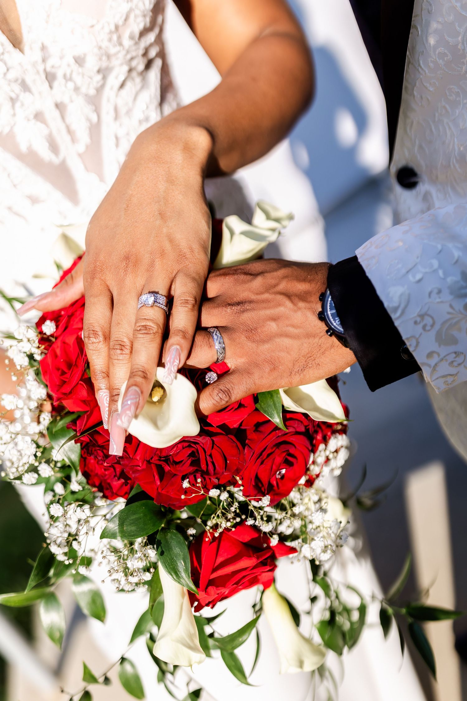 Close-up of hands with wedding rings resting on a bouquet of red roses and white flowers.