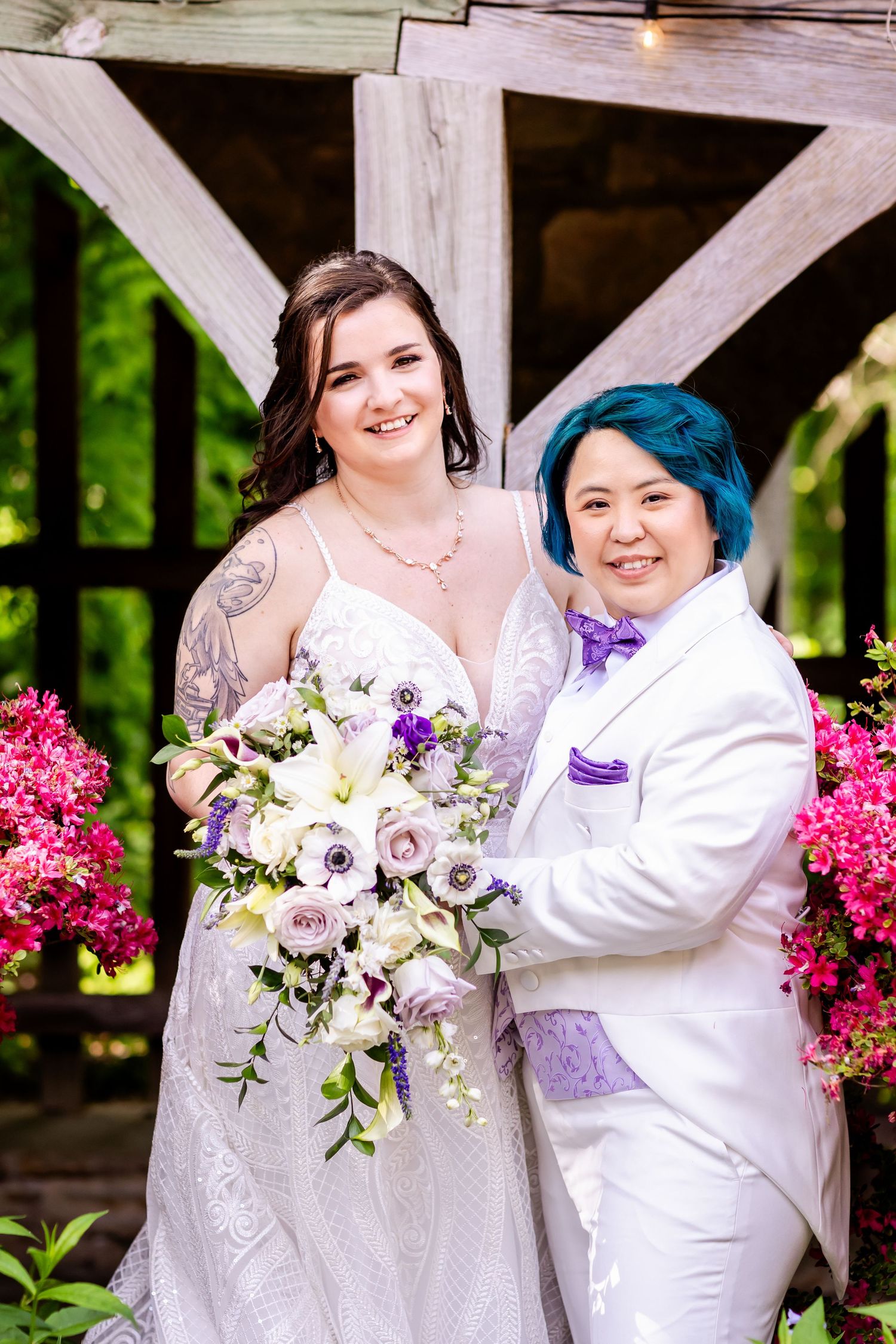 A wedding couple poses together holding a beautiful white and purple floral bouquet.