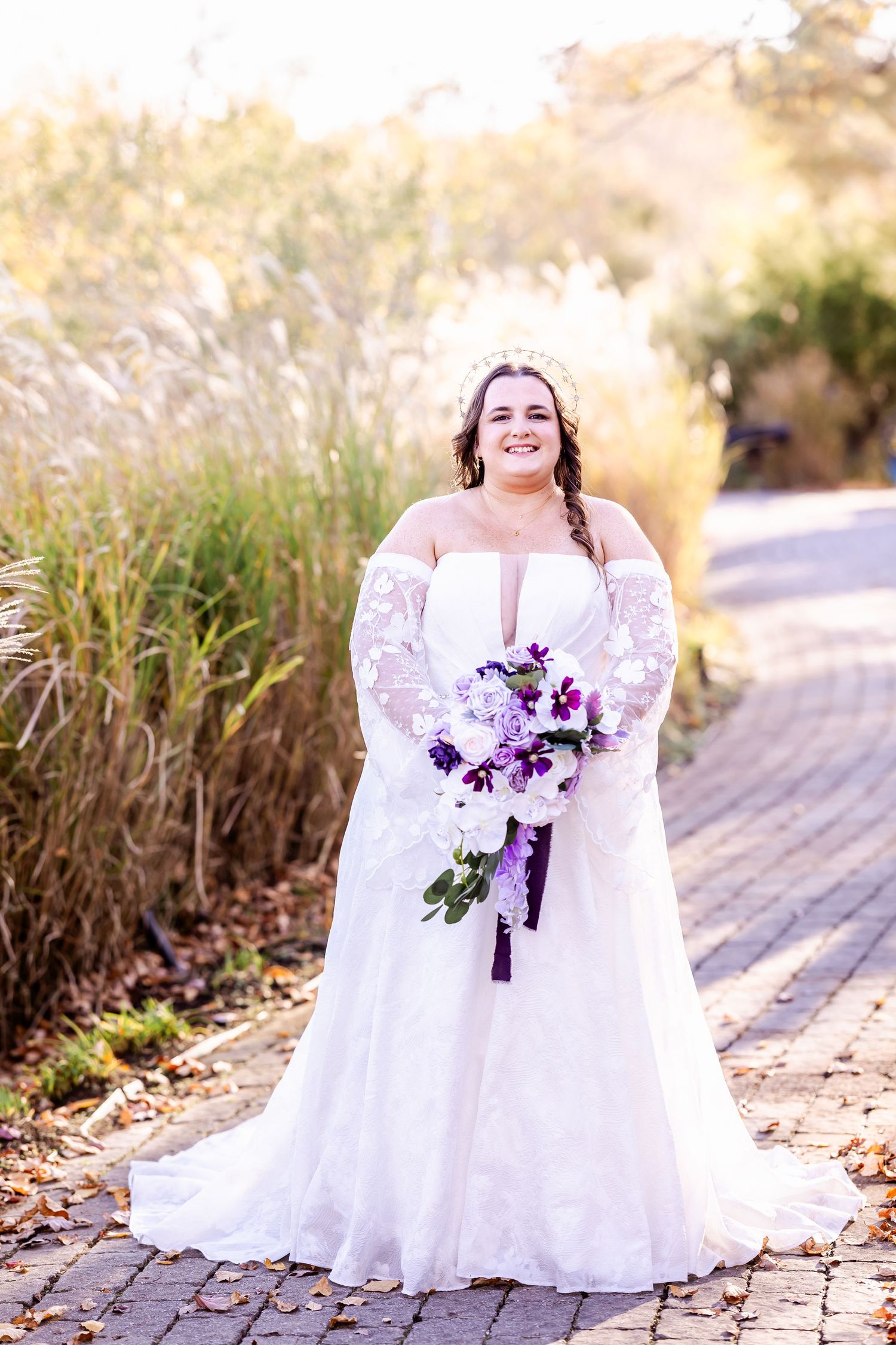 Bridal portrait with off-shoulder lace gown and purple bouquet against natural outdoor setting.