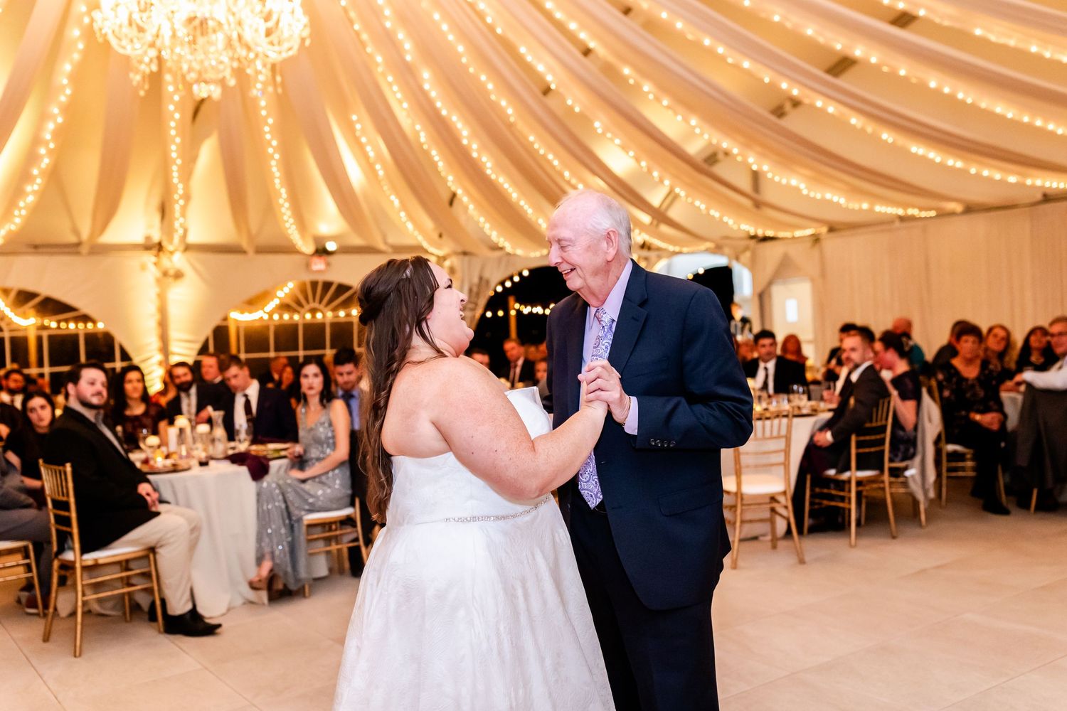 Wedding reception dance in tent venue with elegant draped ceiling and chandelier lighting.