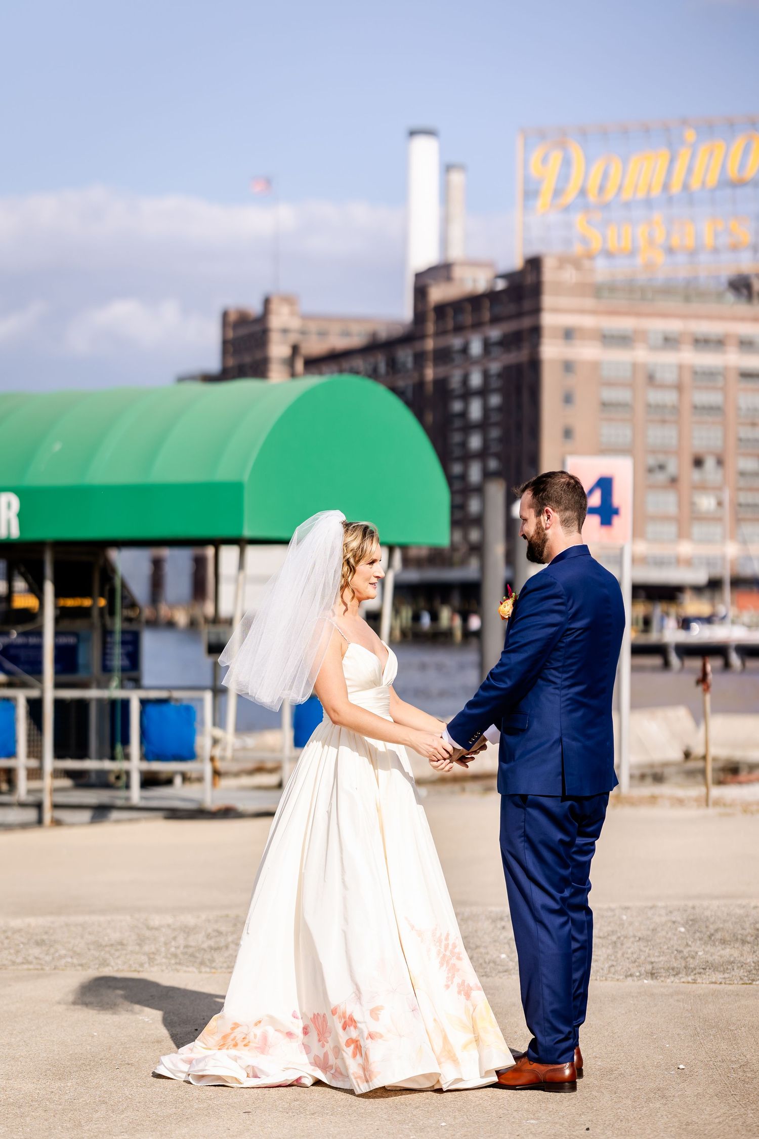 Bride and groom hold hands in front of historic Domino Sugar building with green awning.