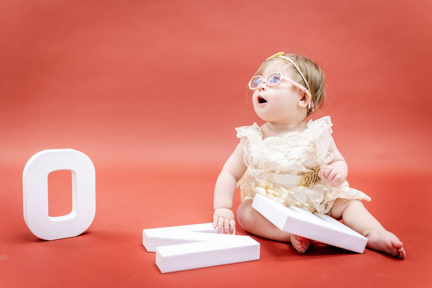 A baby in white dress sits next to decorative letters on a red backdrop.
