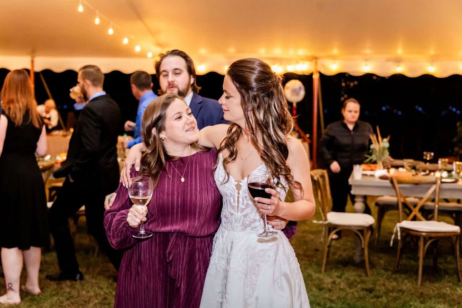 Wedding guests laugh and dance together under a tent with twinkling lights at dusk.