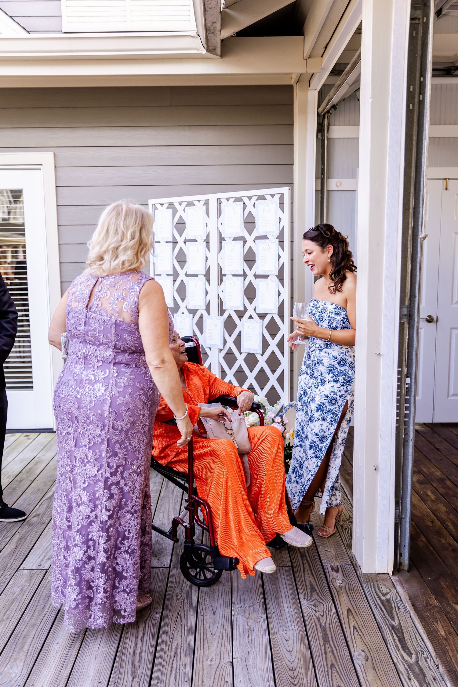 Wedding guests in formal attire gather on a wooden deck outside a gray house.