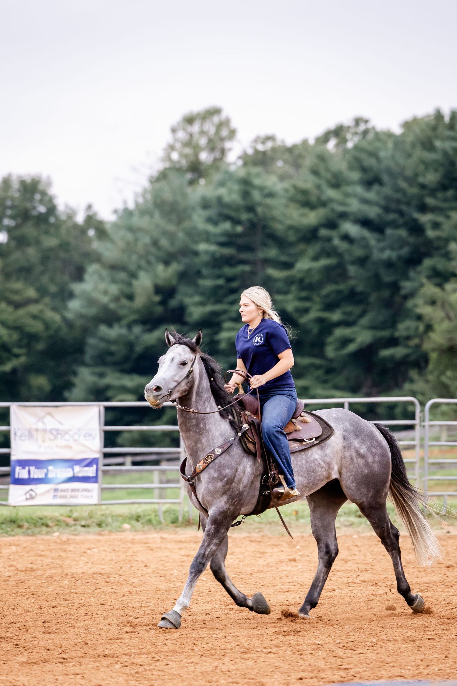 Rider practicing barrel racing pattern on grey horse in outdoor dirt arena with pine trees in background.