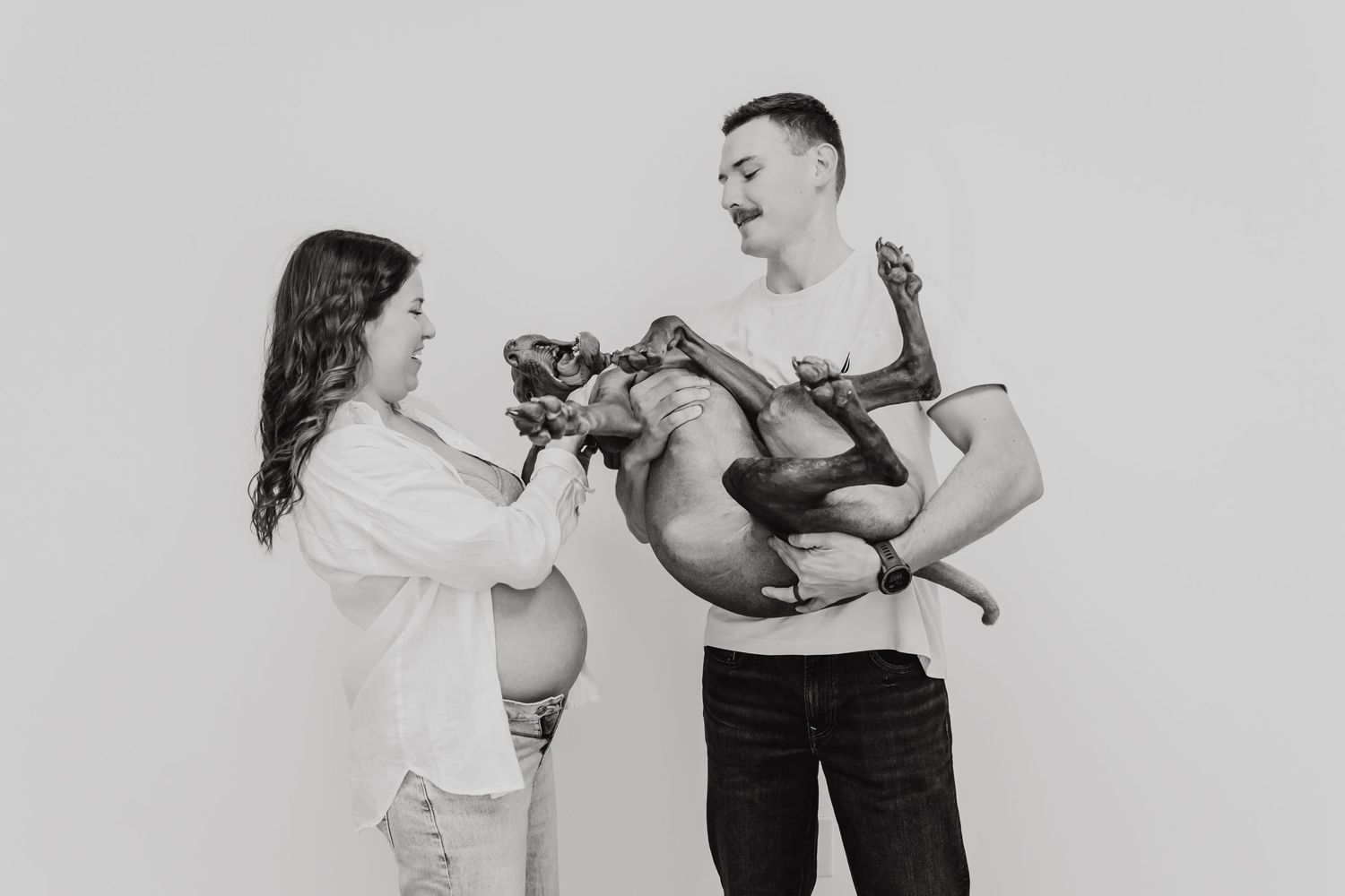 Black and white photo of a couple laughing while holding their French Bulldog puppy against white background.