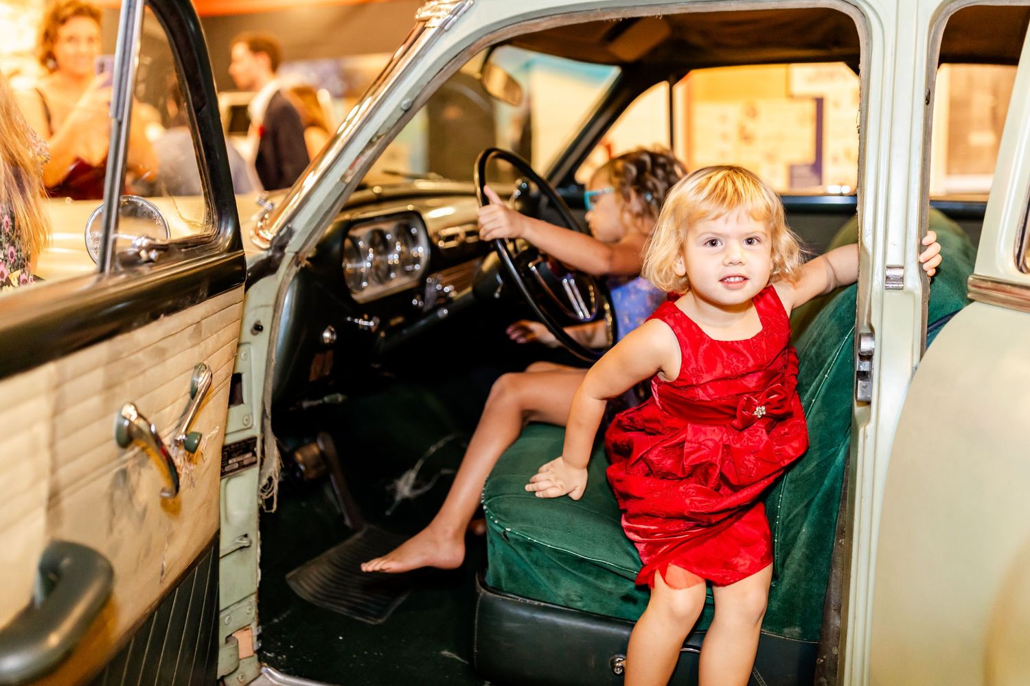 A small child in a red dress sits in the front seat of a vintage automobile.
