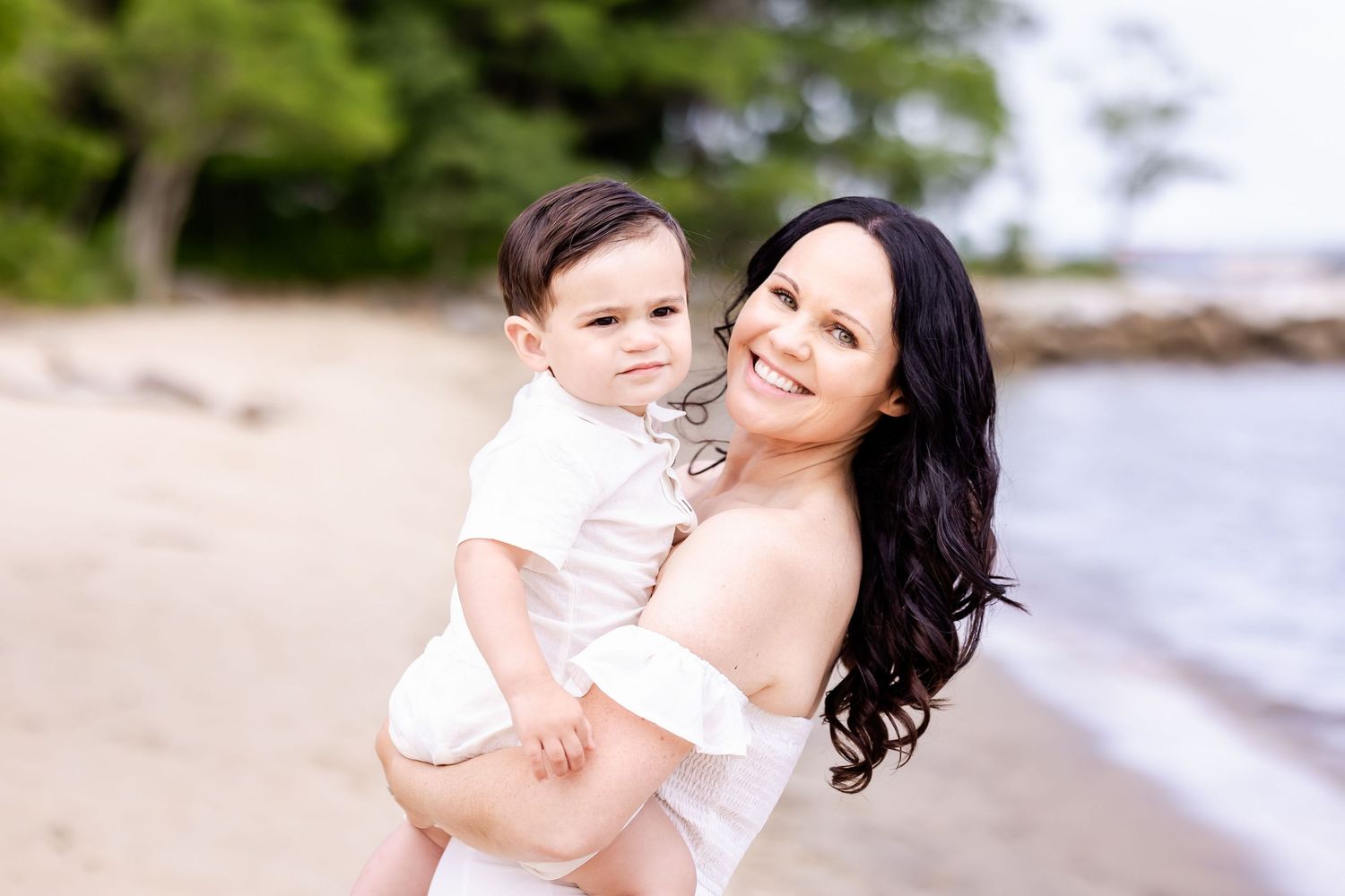 A tender moment between a parent and child on a scenic beach with water and trees in the background.