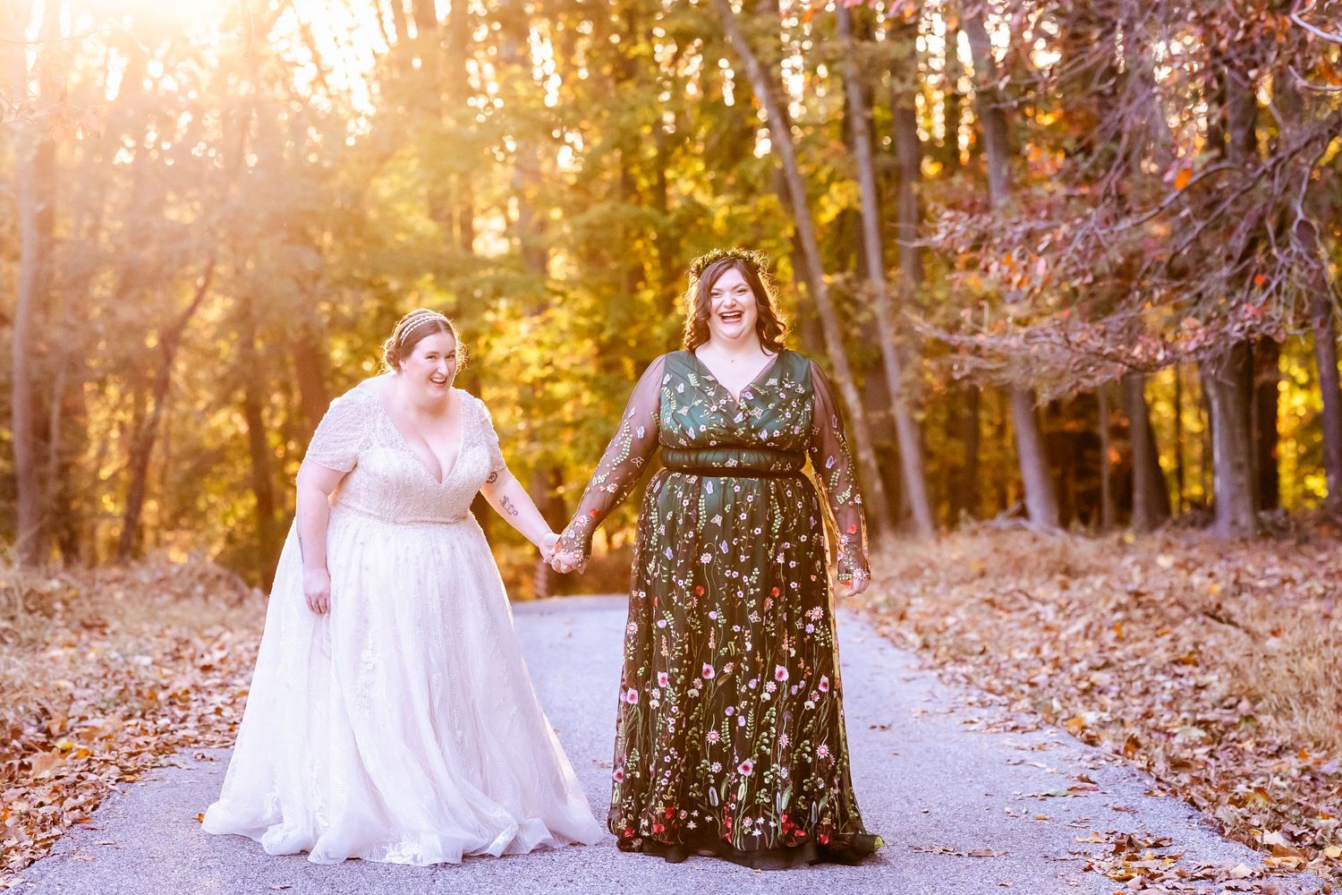 Two people in elegant formal dresses hold hands on an autumn path at sunset with golden light filtering through trees.