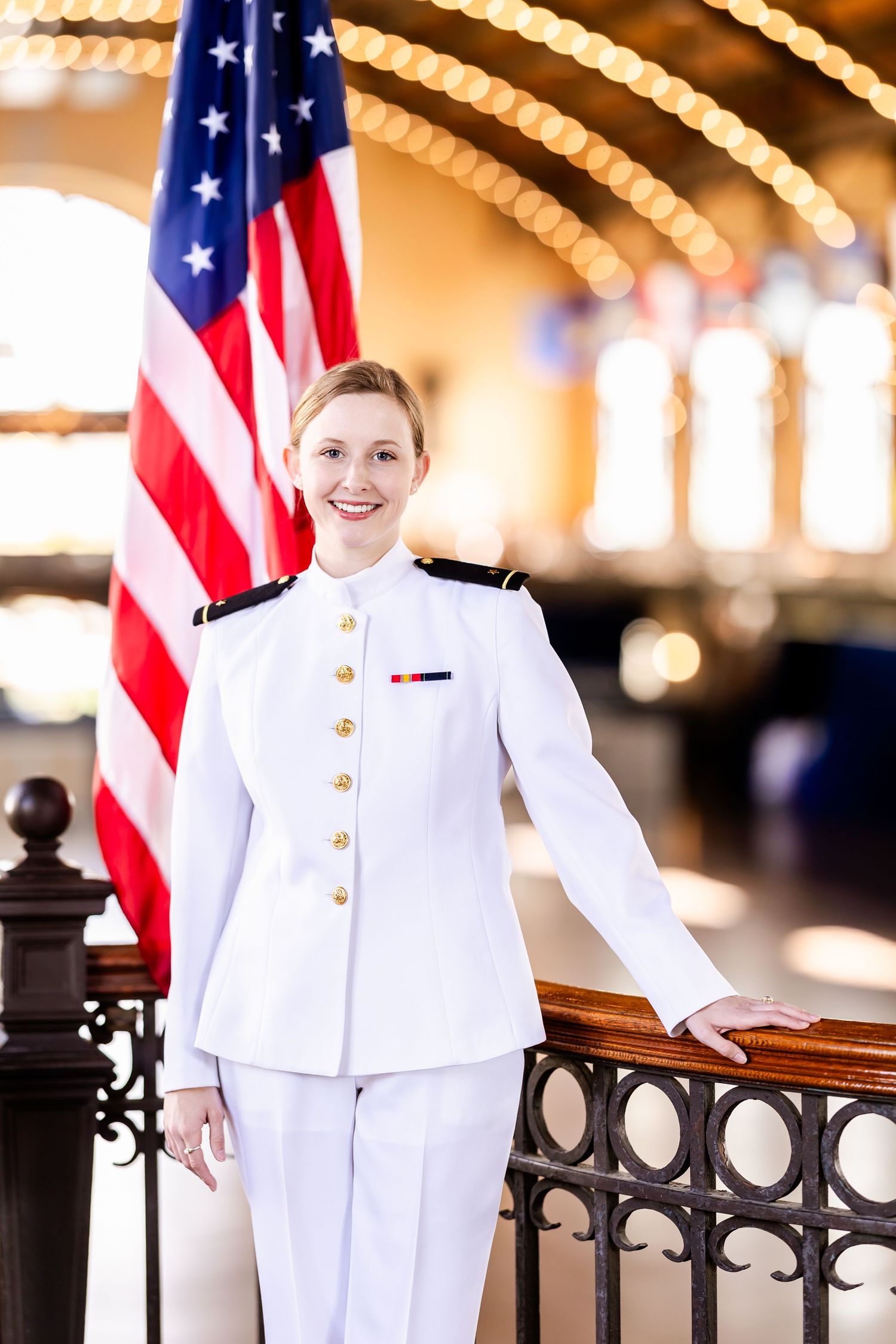 Naval officer in white dress uniform standing at attention with American flag in formal setting.