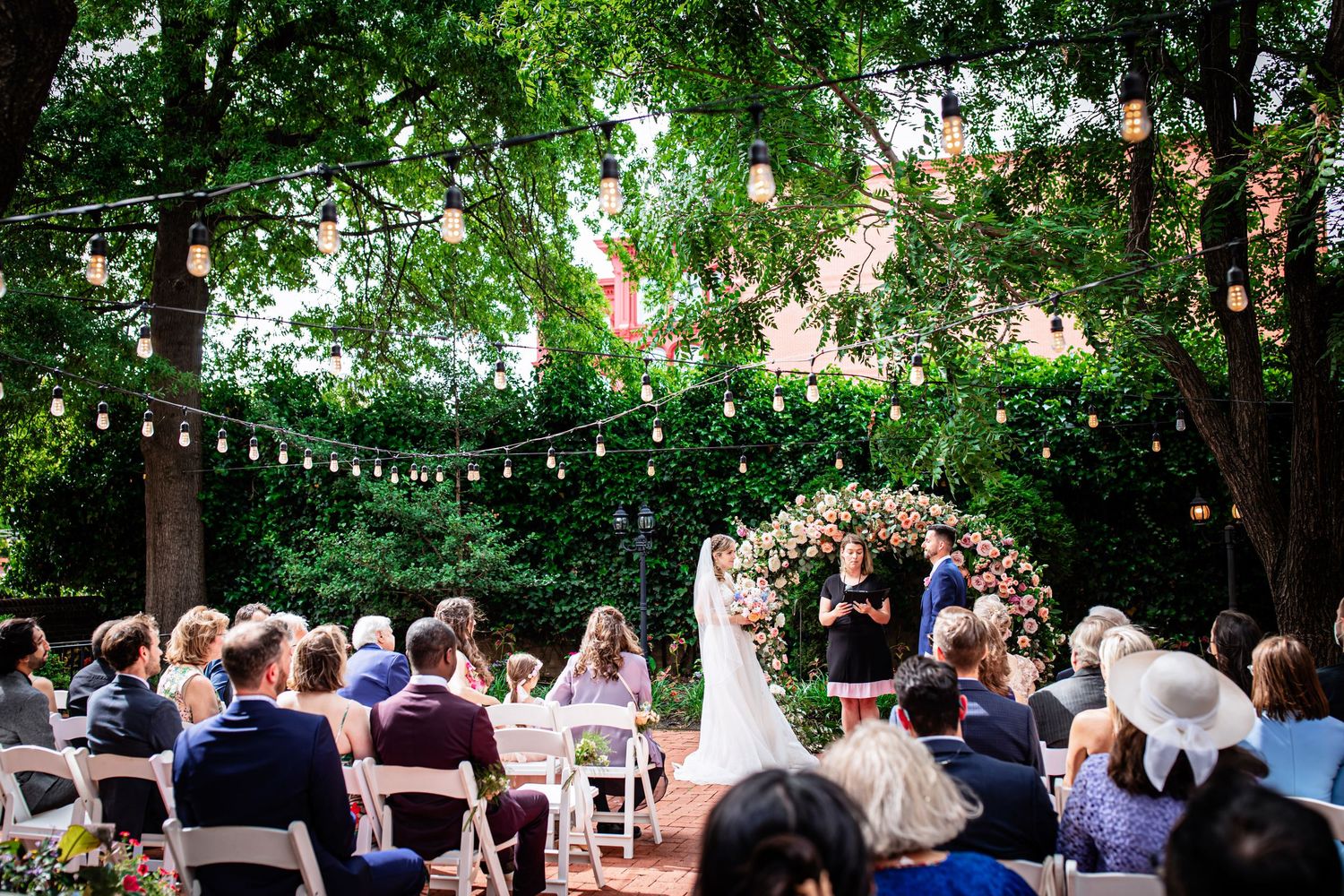 Outdoor garden wedding ceremony under string lights with guests seated in white chairs beneath leafy trees.