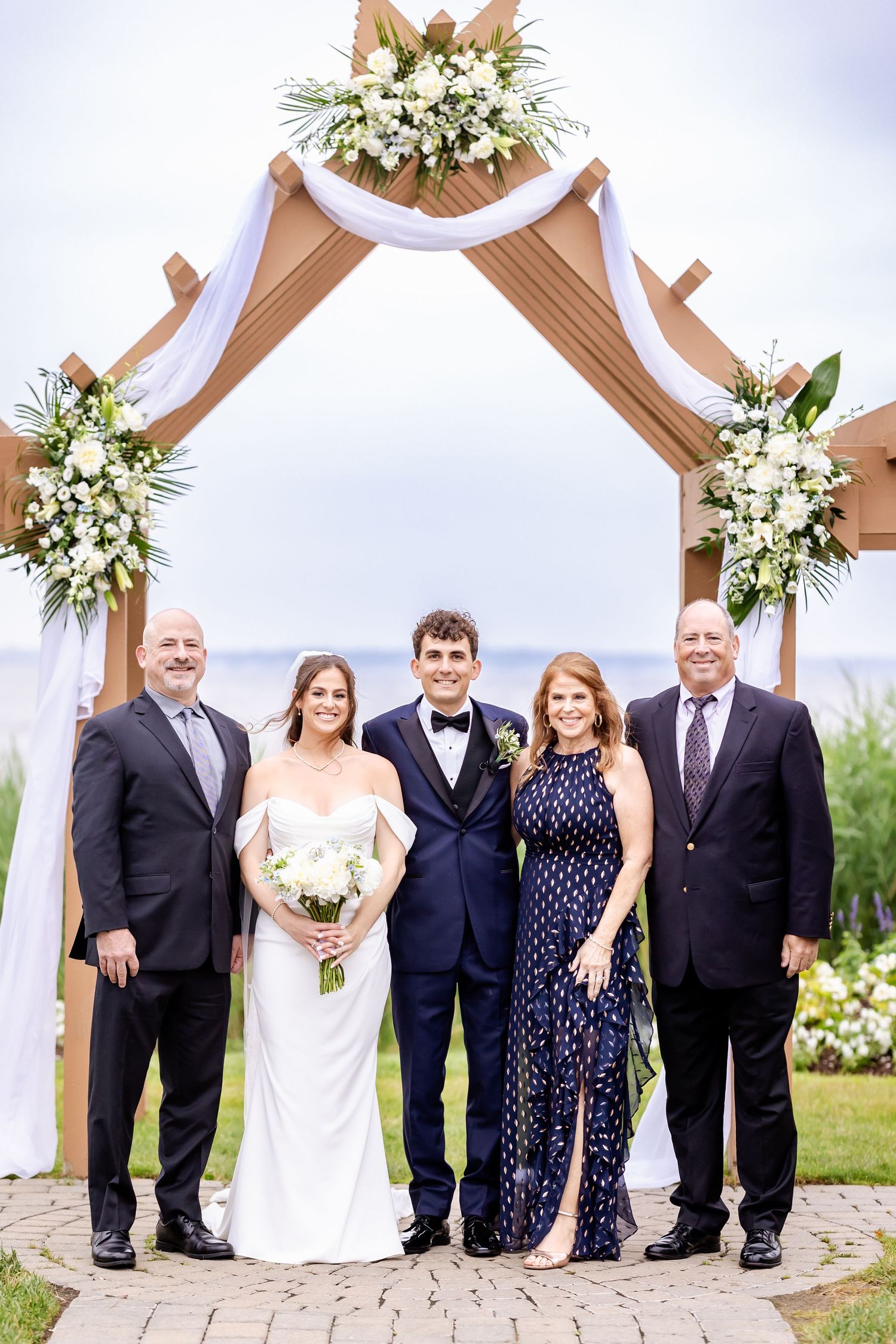 Wedding party poses under wooden pergola decorated with white floral arrangements and draping.