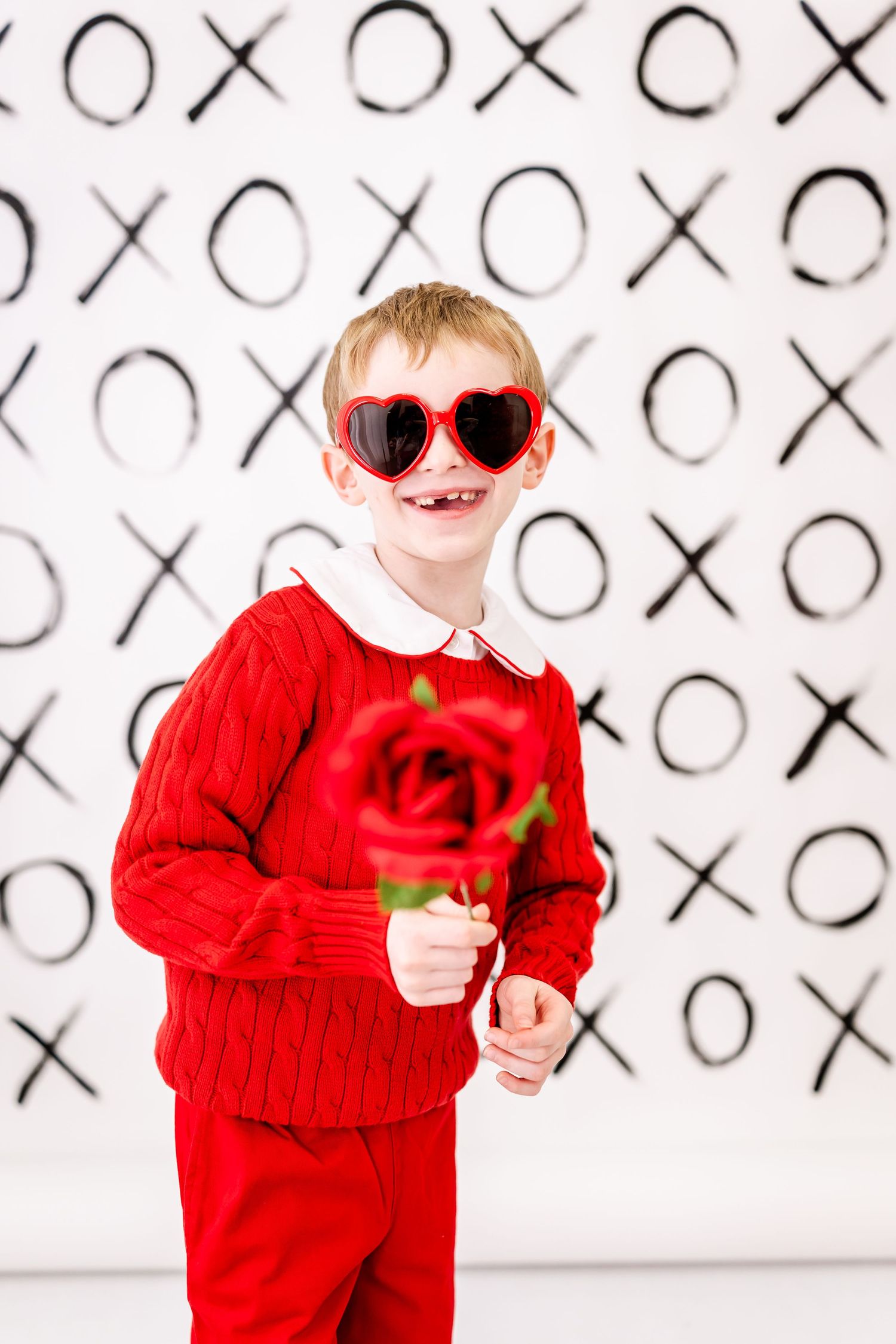 A child in red festive attire wearing heart sunglasses holds a rose against a black and white X's and O's background.