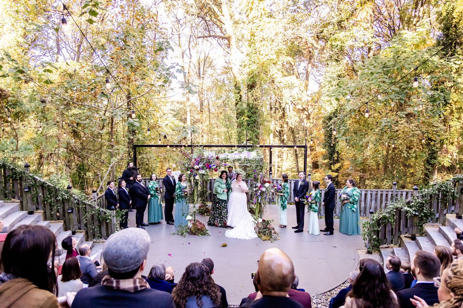 An outdoor wedding ceremony takes place on a tree-lined pathway with guests seated on either side during autumn.