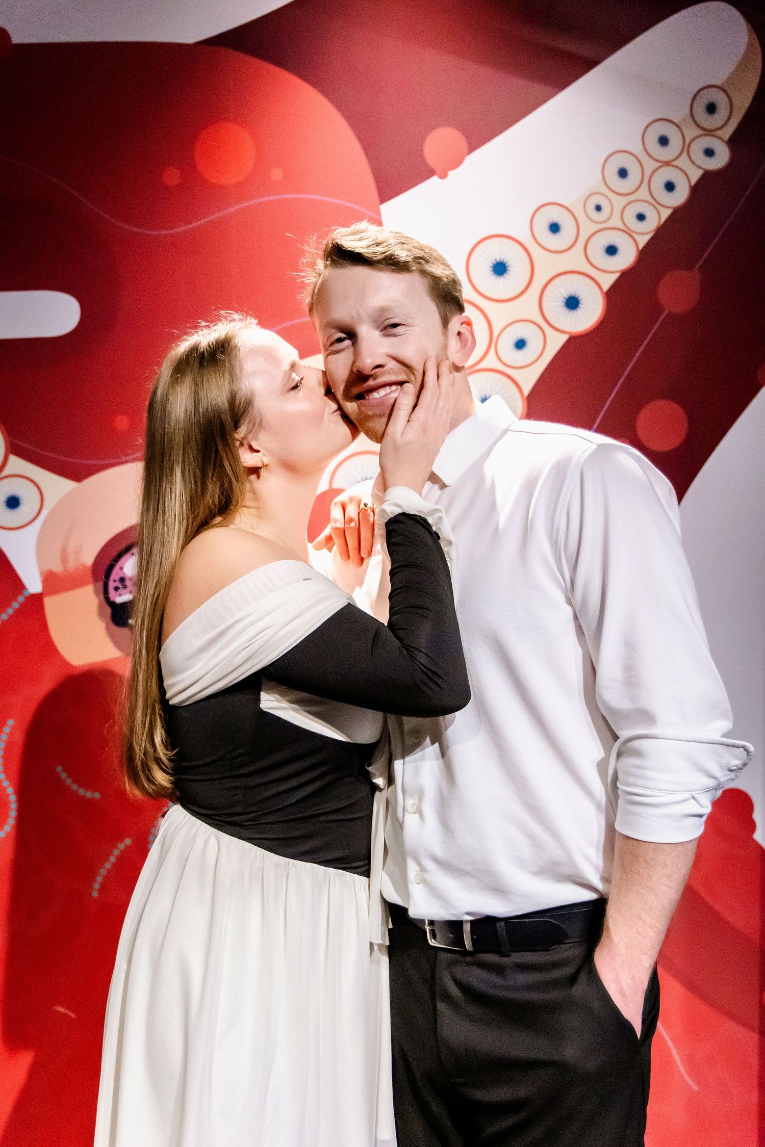 A couple shares an affectionate moment against a red and white patterned backdrop.