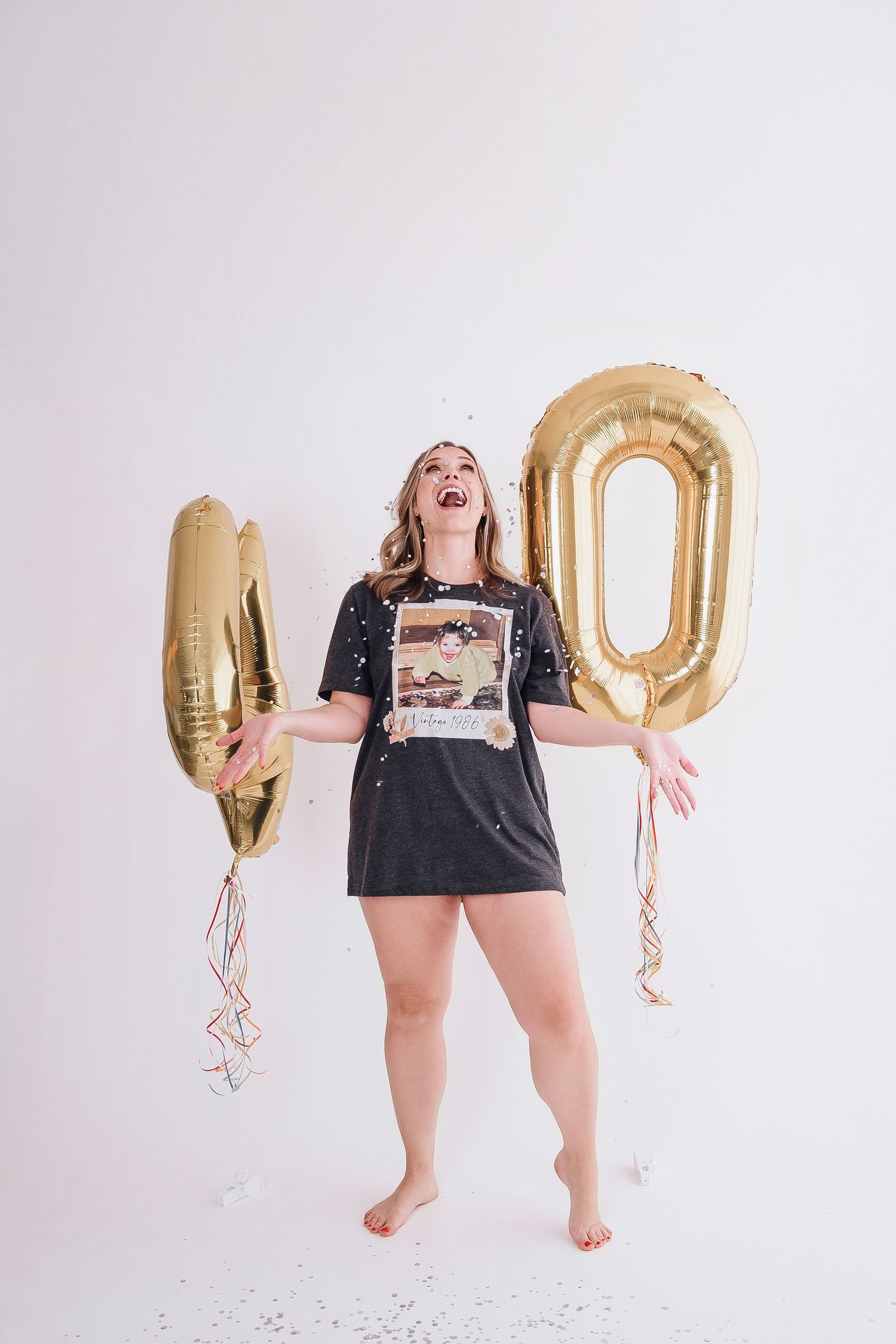 Woman in black shirt jumps playfully with large gold number 40 balloons against white background.
