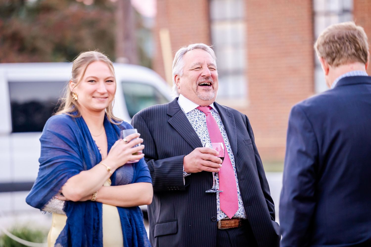 Two guests laughing and enjoying drinks at an outdoor wedding reception, woman in blue shawl and man in pink tie.