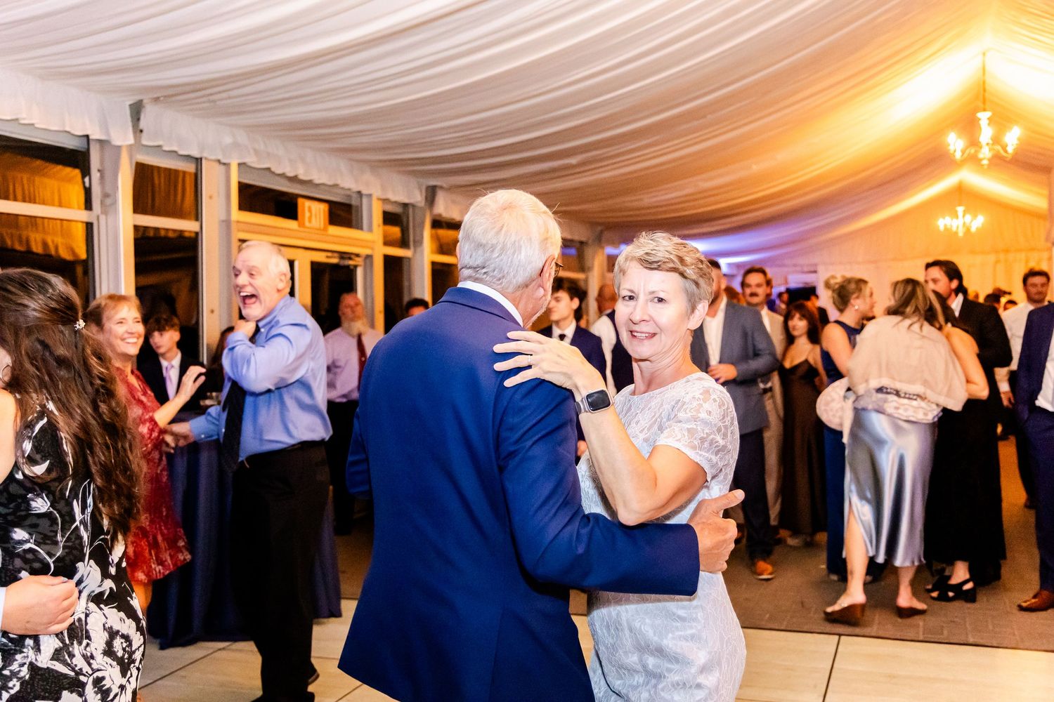 Wedding celebration with guests dancing under warm lighting and white tent draping.