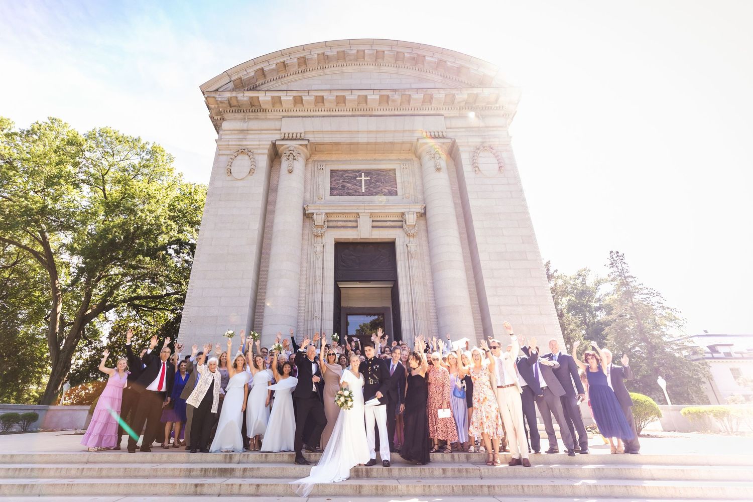 Large wedding party poses on grand marble steps of a neoclassical building with columns and domed roof in sunlight.