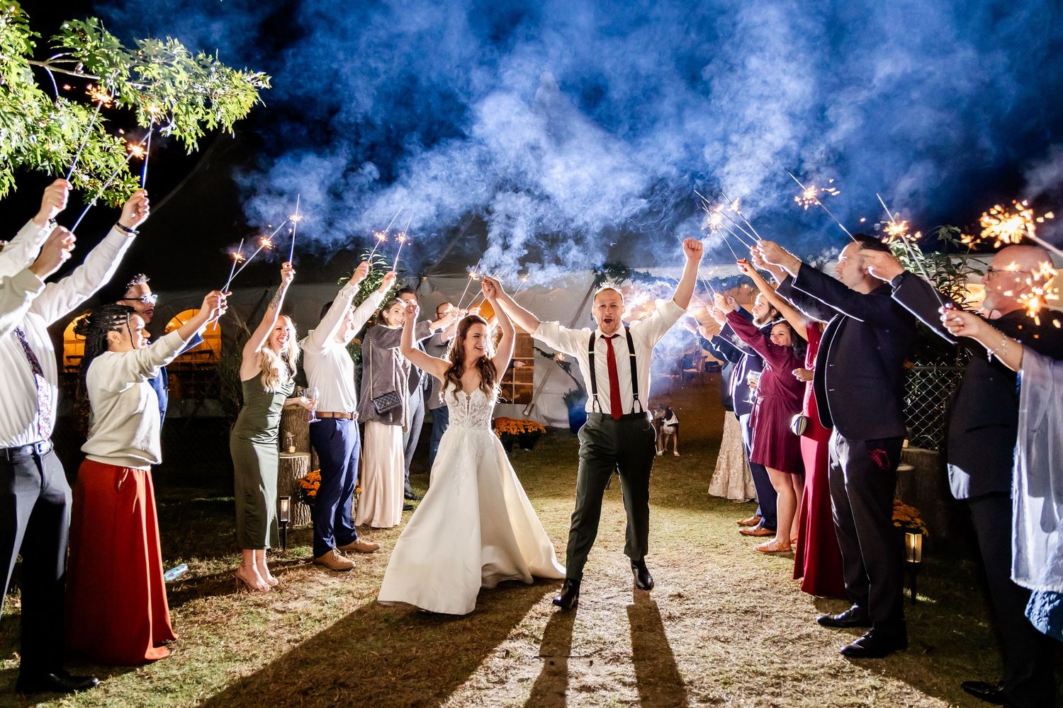 Wedding guests creating a sparkler exit tunnel outdoors with blue smoke in the background.
