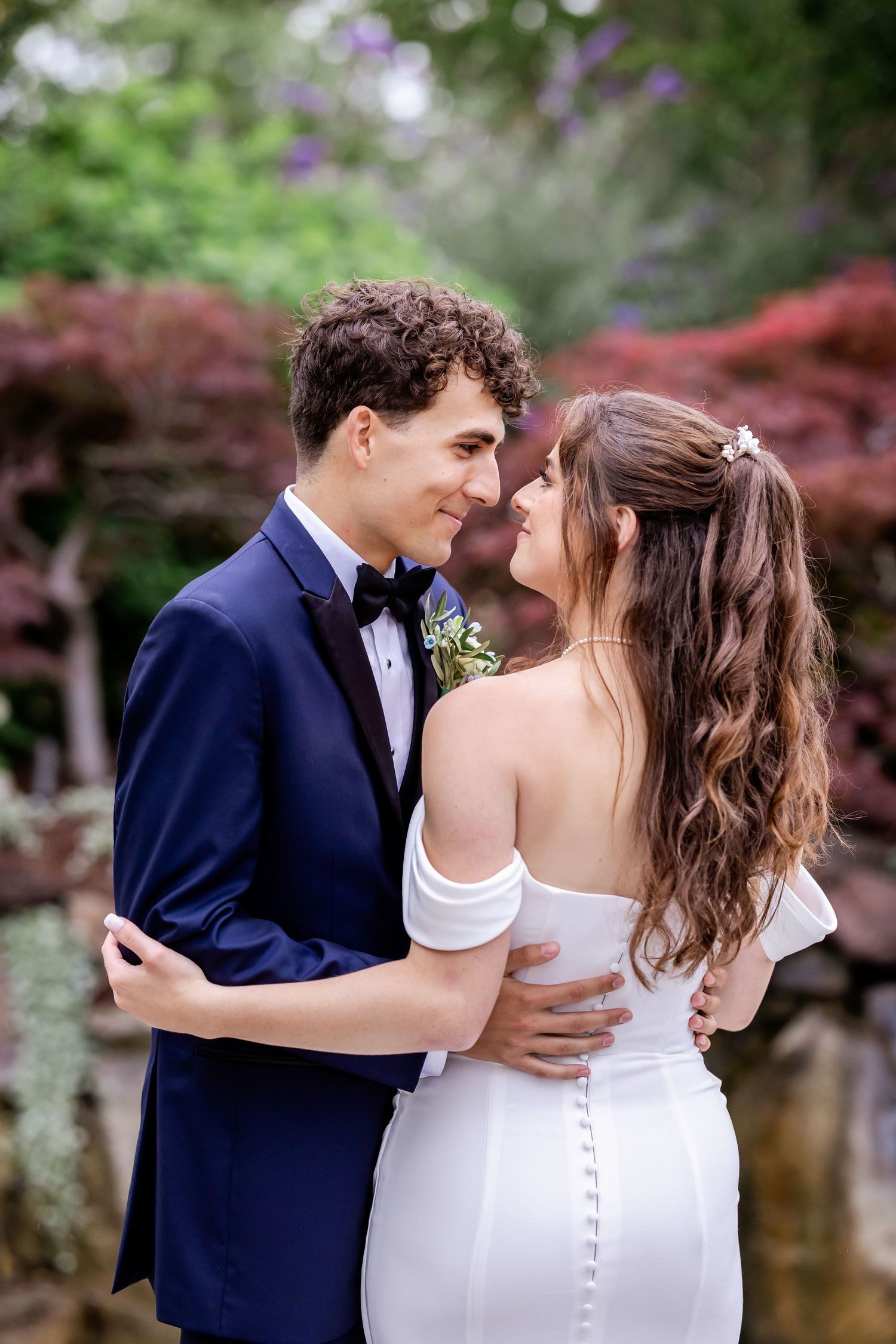 A romantic wedding couple shares intimate moments in a garden with purple flowers and red maples.