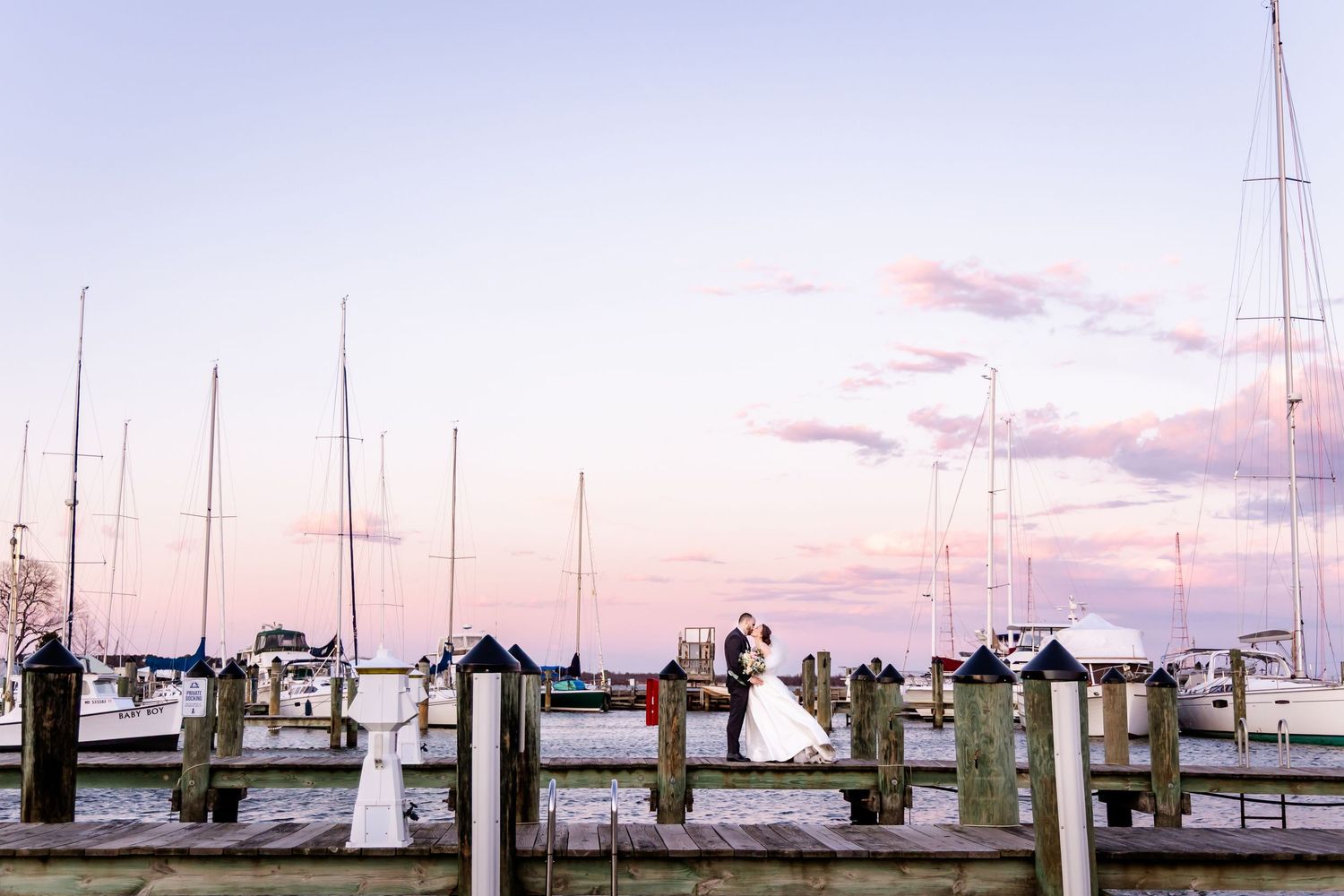 Wedding couple stands on wooden dock at dusk with sailboats and pink sky creating dreamy waterfront scene.