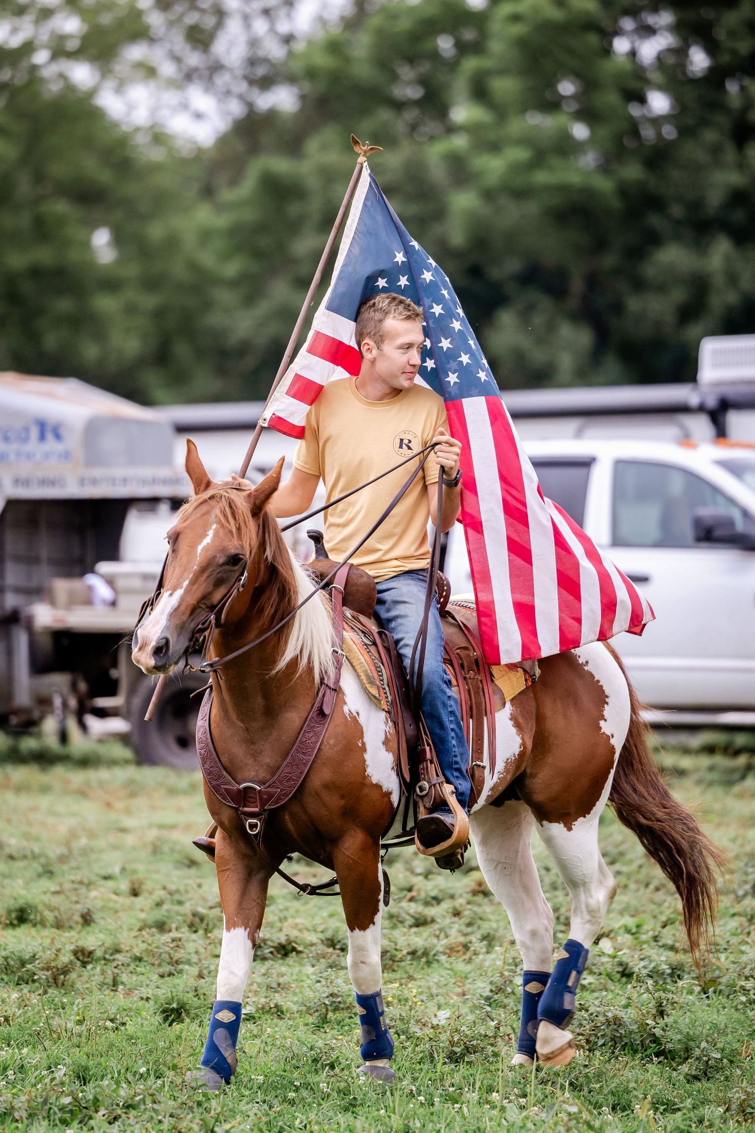 Rider carrying American flag on horseback with protective boots in grassy field.
