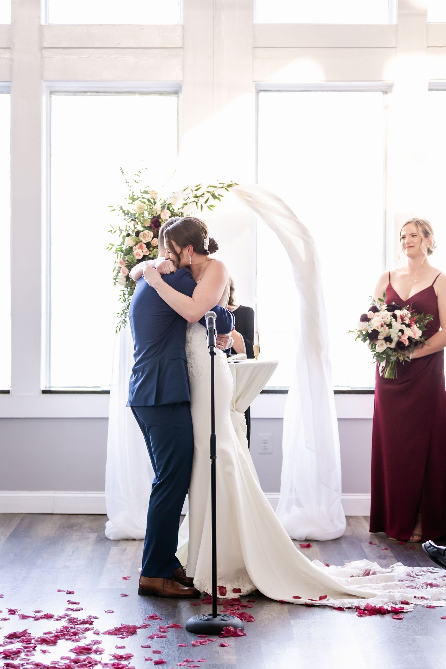 Wedding couple and attendants in burgundy dresses standing in a bright white room with rose petals on the floor.