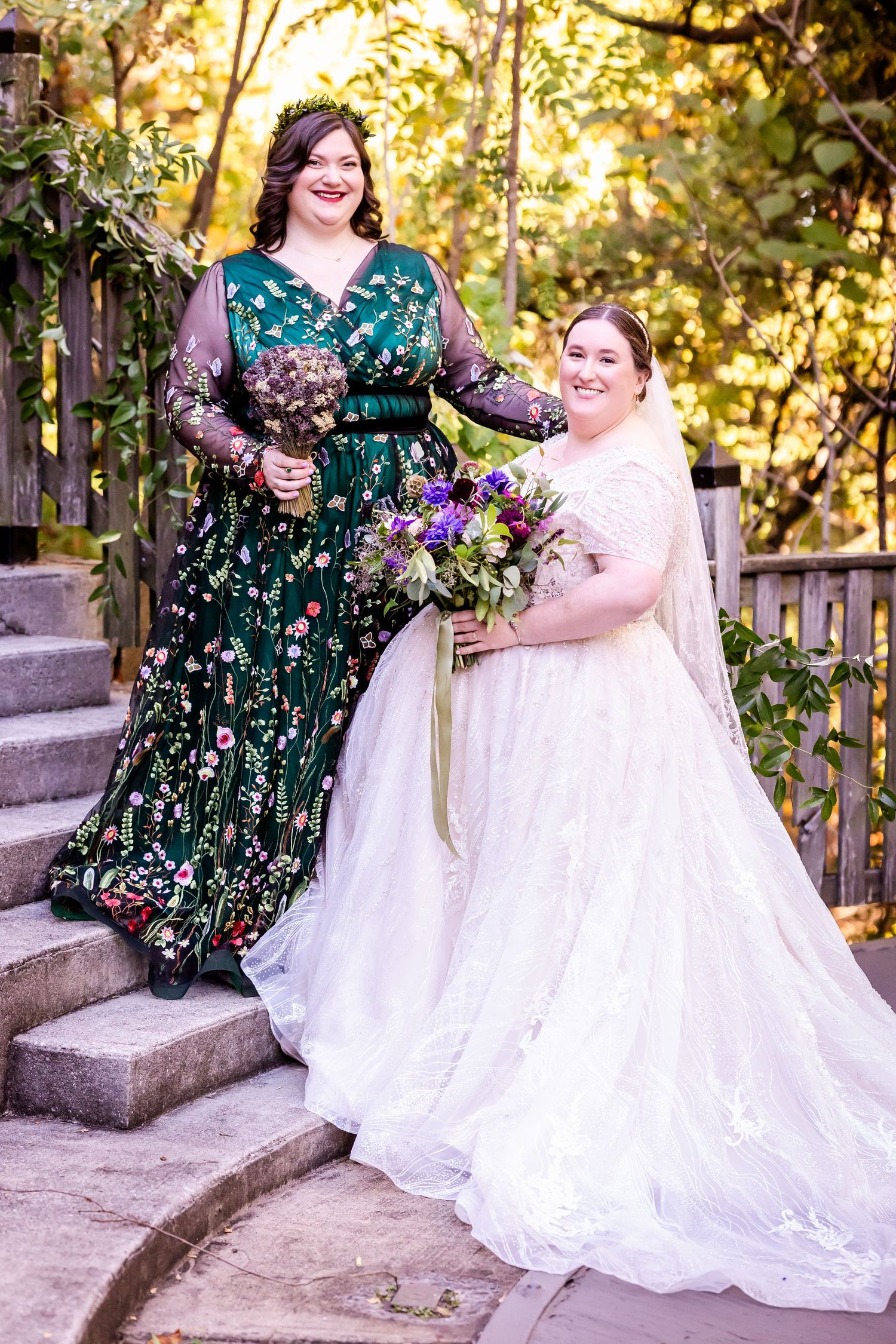 Two brides pose on stone steps, one wearing a floral green dress and one in traditional white.