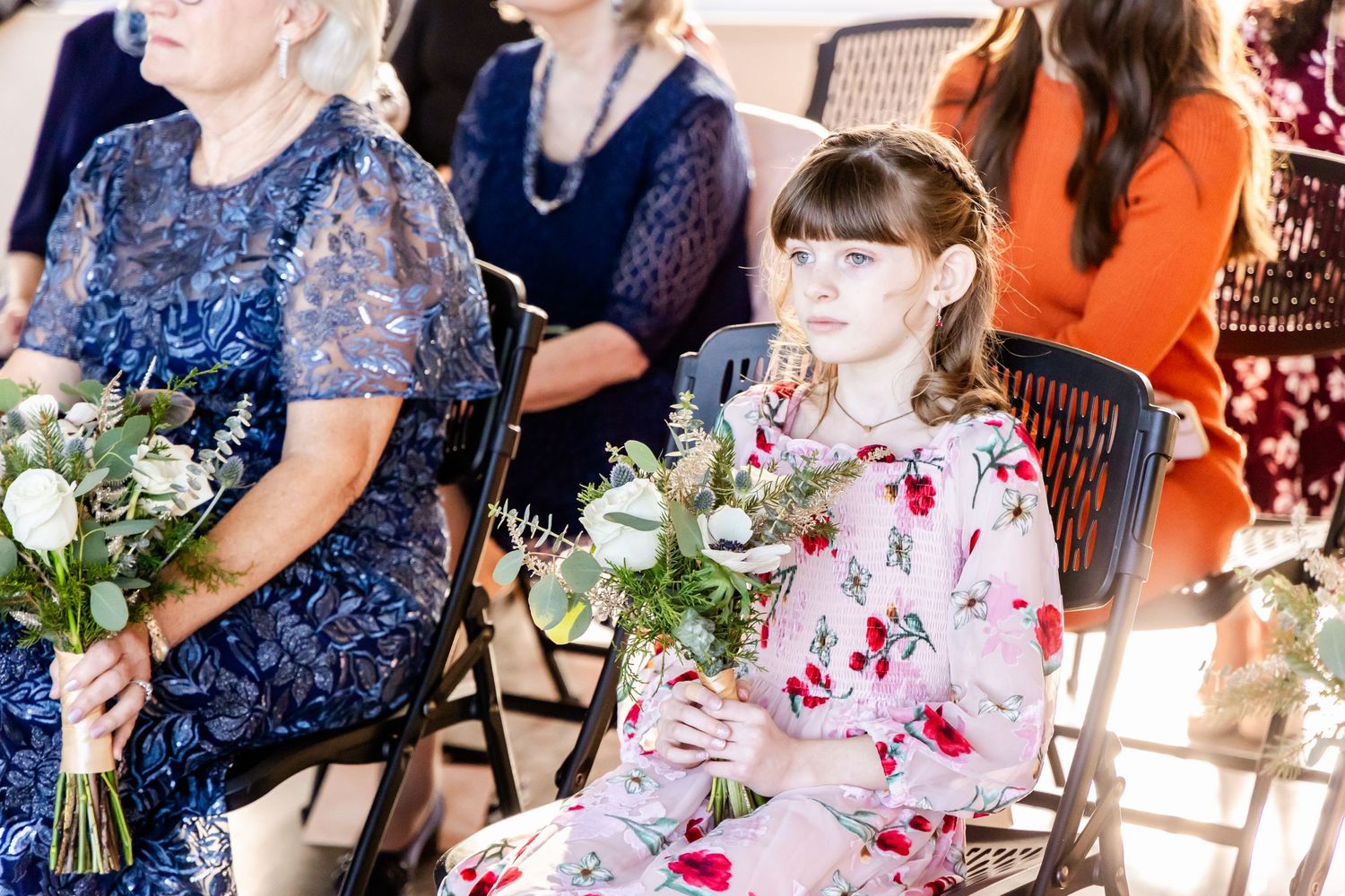Two women in dresses holding bouquets and sitting on chairs during a ceremony.