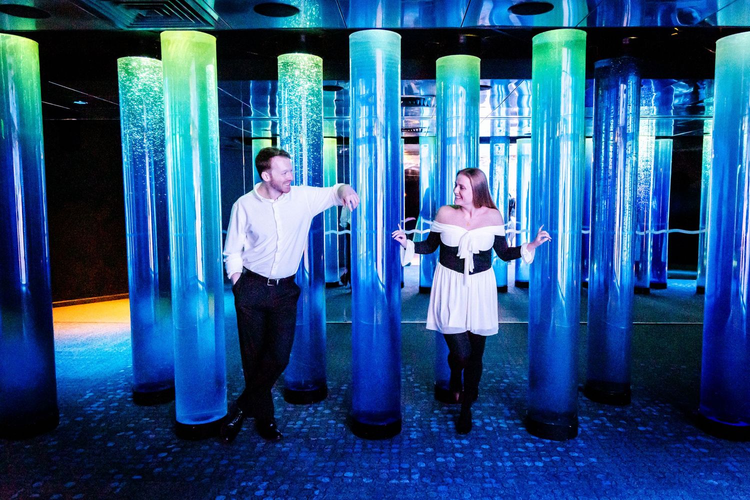 Two people interact with glowing blue cylindrical displays in a dark exhibition space.