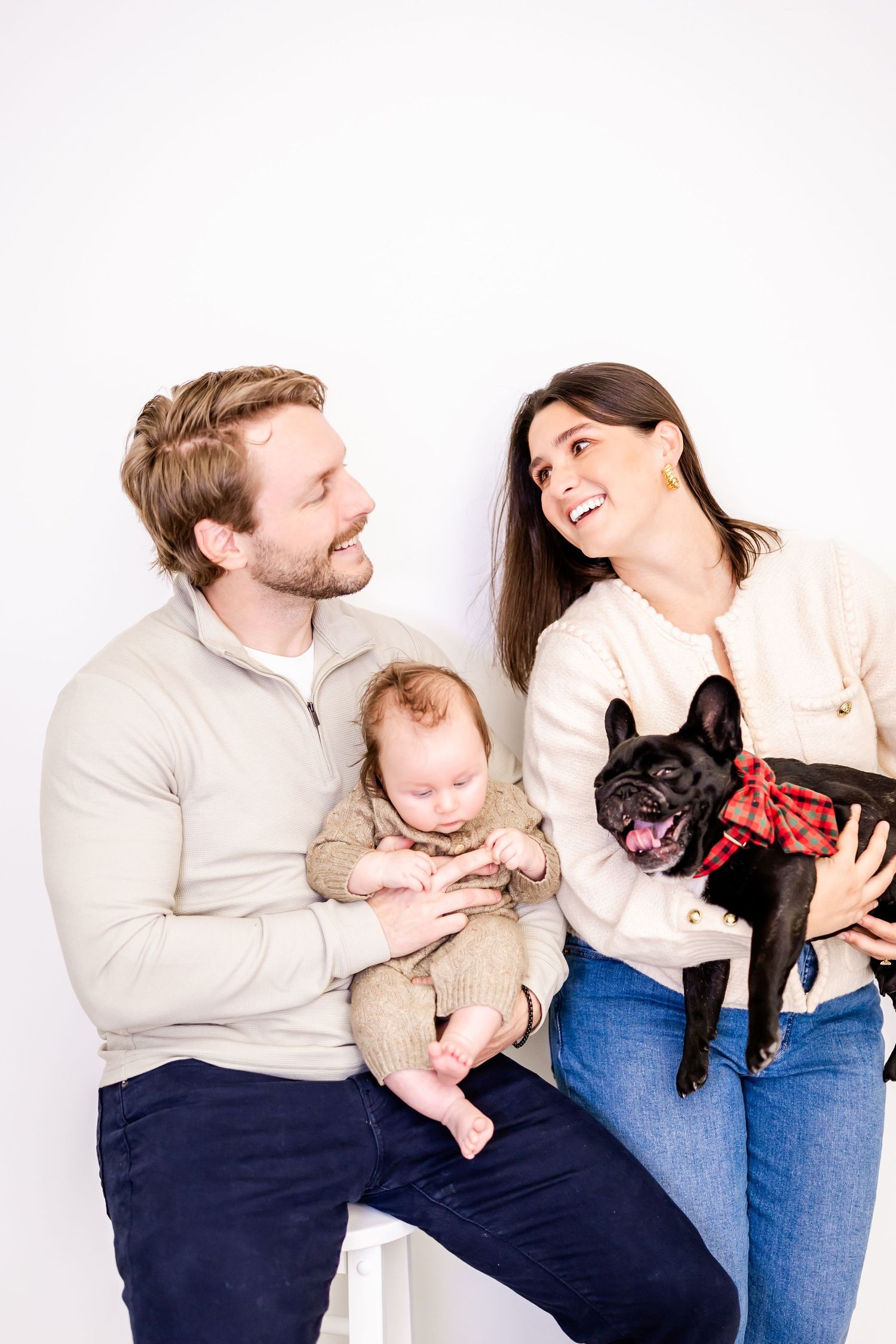 Happy family poses together with their adorable baby and smiling black French Bulldog wearing a red bandana.
