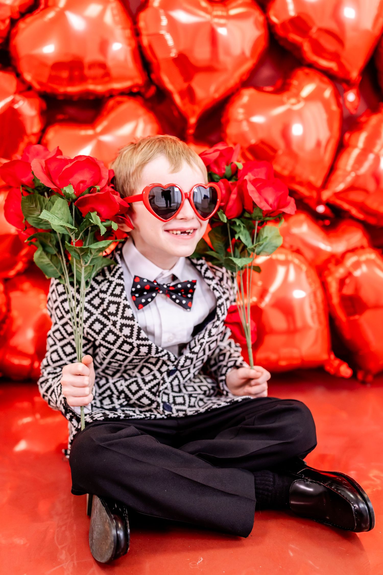 A child in a patterned blazer and heart sunglasses poses with roses against a backdrop of red heart-shaped balloons.