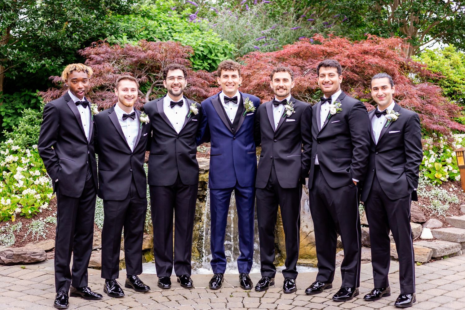 Groomsmen in black tuxedos and bow ties stand together for formal wedding photos in a garden setting.