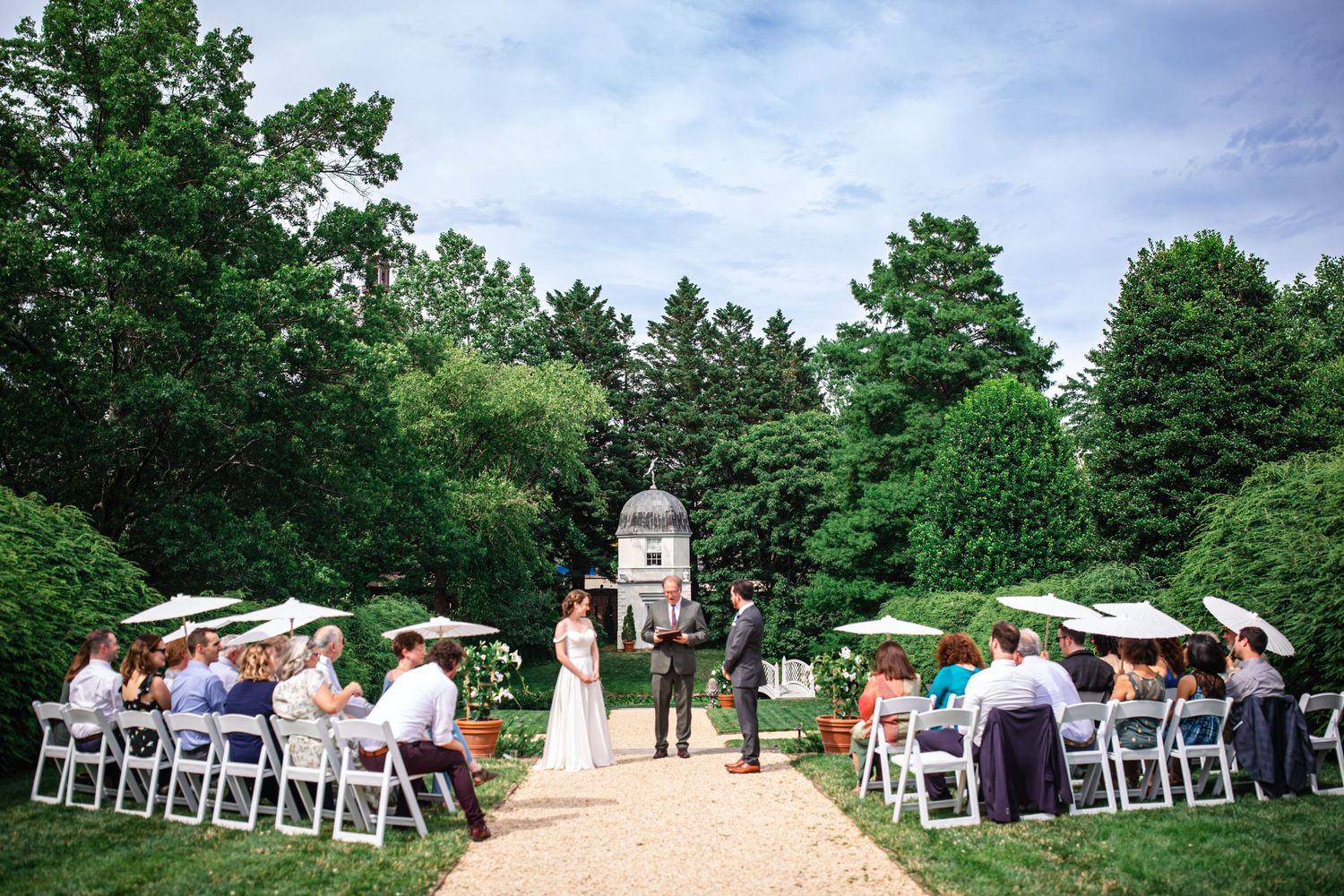 White chairs arranged in rows for an outdoor wedding ceremony with green trees and blue sky in the background.