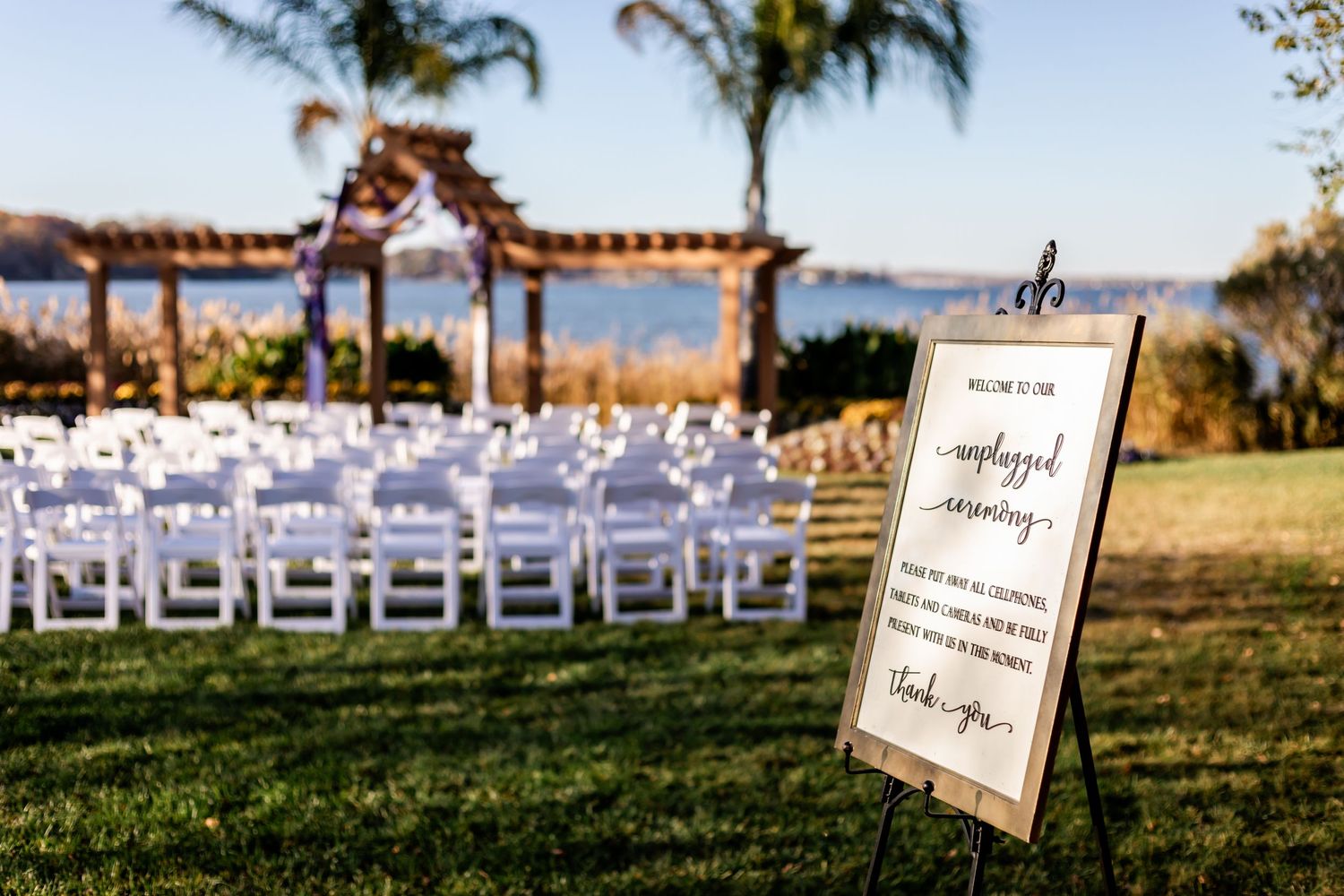 Outdoor wedding ceremony setup with white chairs and wooden arch overlooking waterfront view.