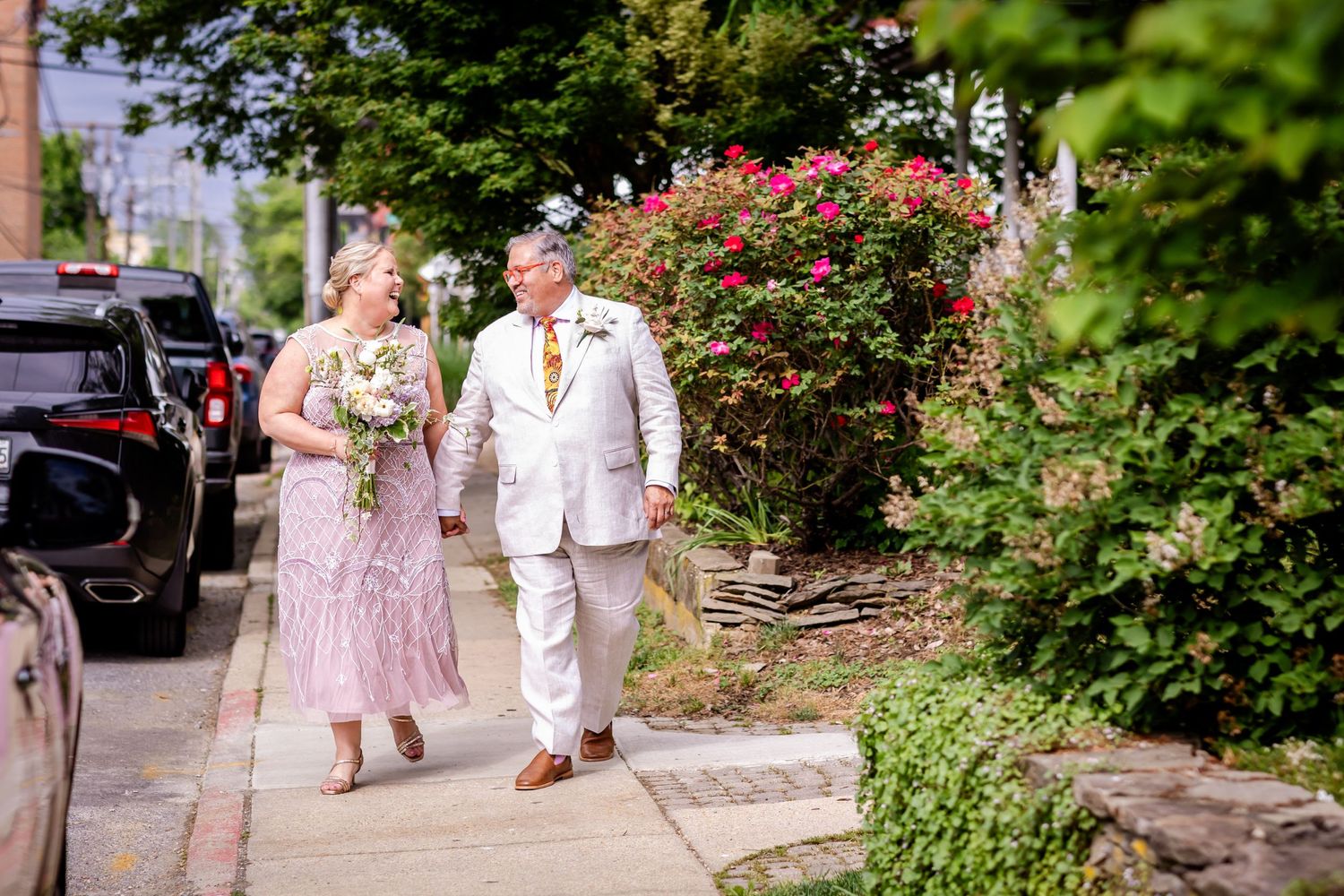 A couple walks along a garden path in formal attire with blooming flowers in the background.