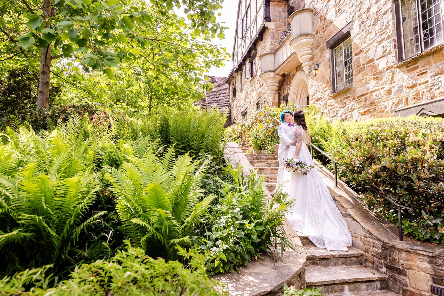 A romantic wedding photo on stone steps leading to an old stone building surrounded by ferns and gardens.
