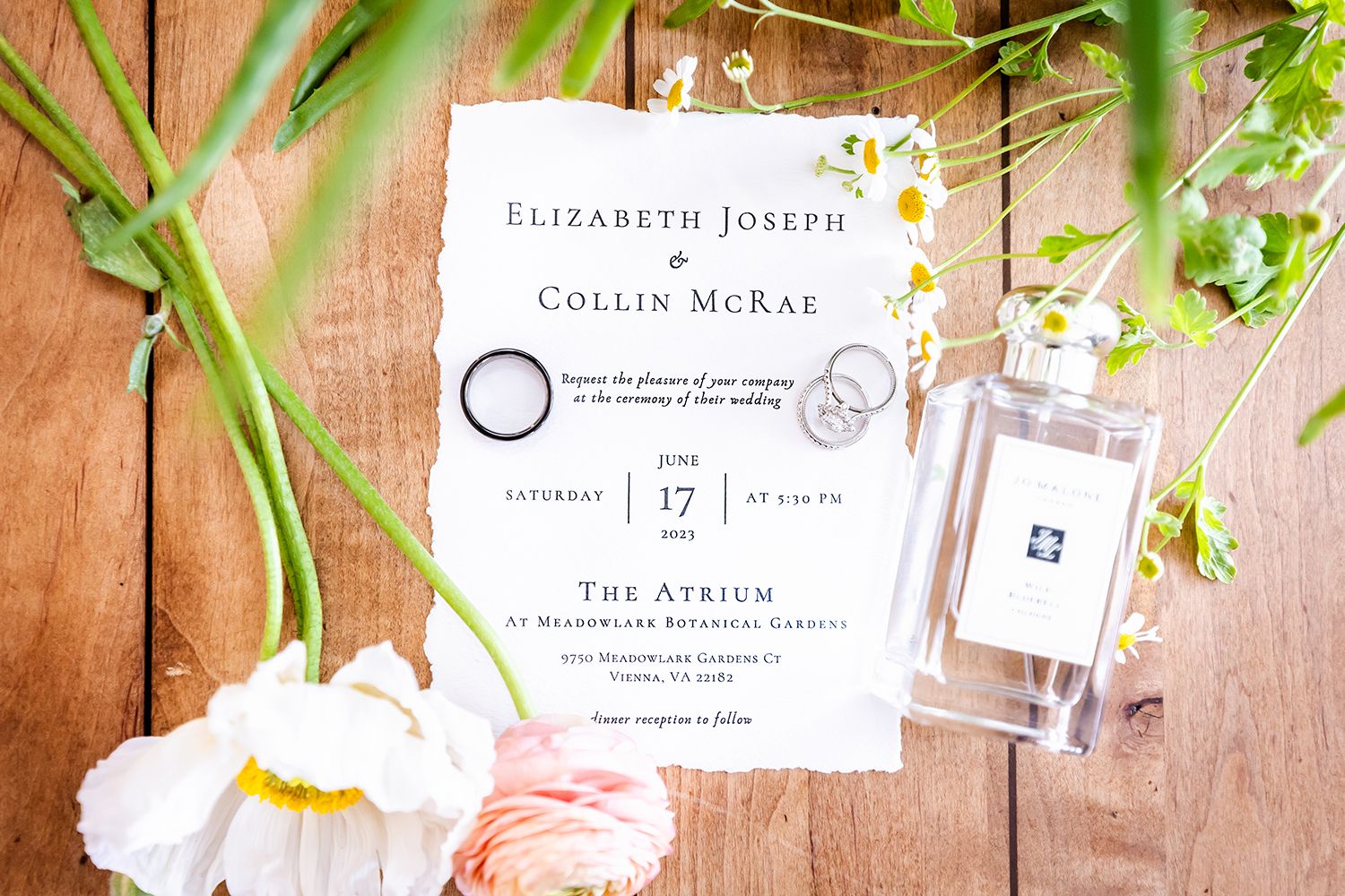 An elegant white wedding invitation displayed with pink tulips and perfume bottle on rustic wooden background.