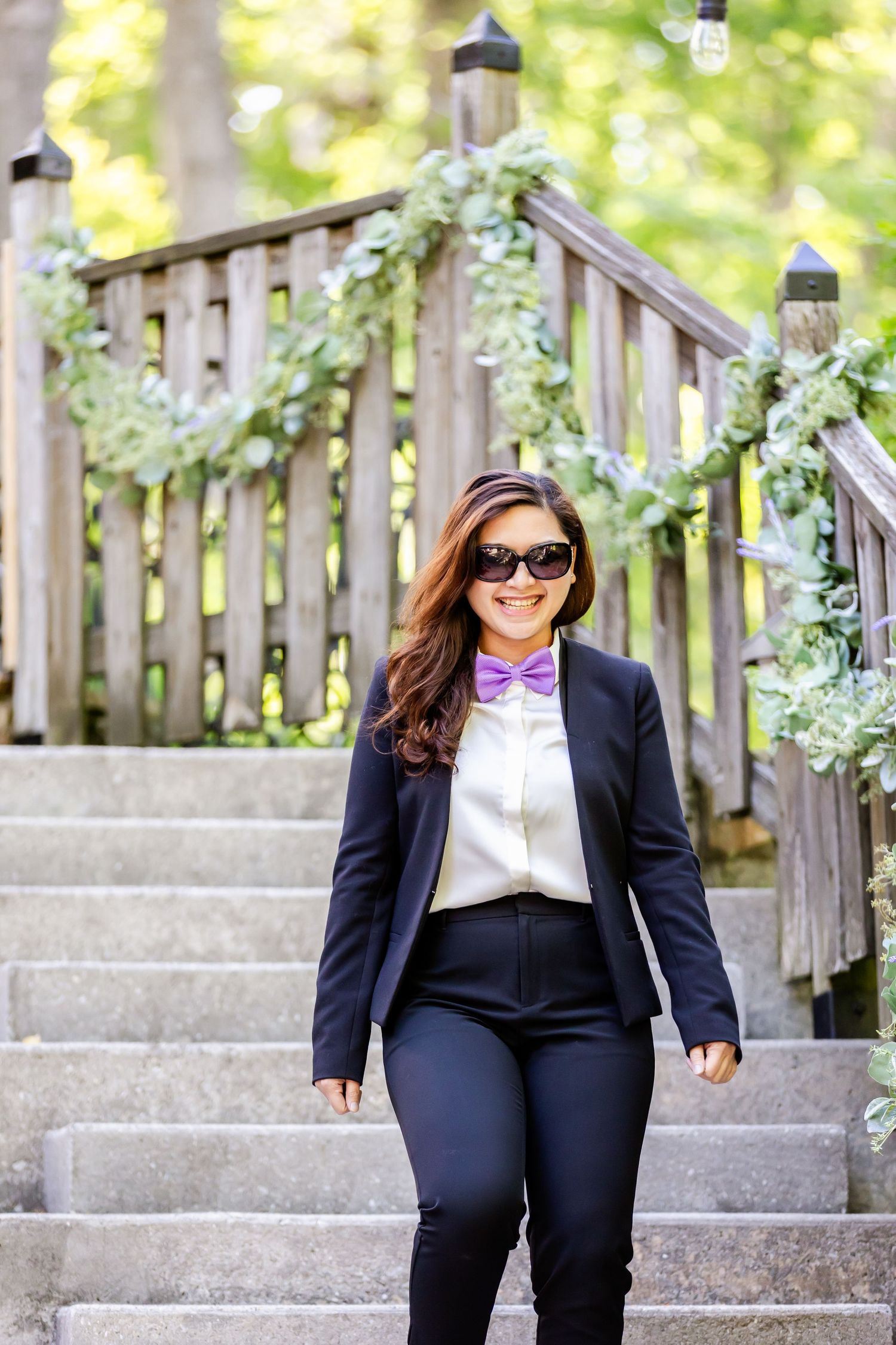 Stylish woman in business suit with purple bowtie descends garden stairs decorated with floral greenery.