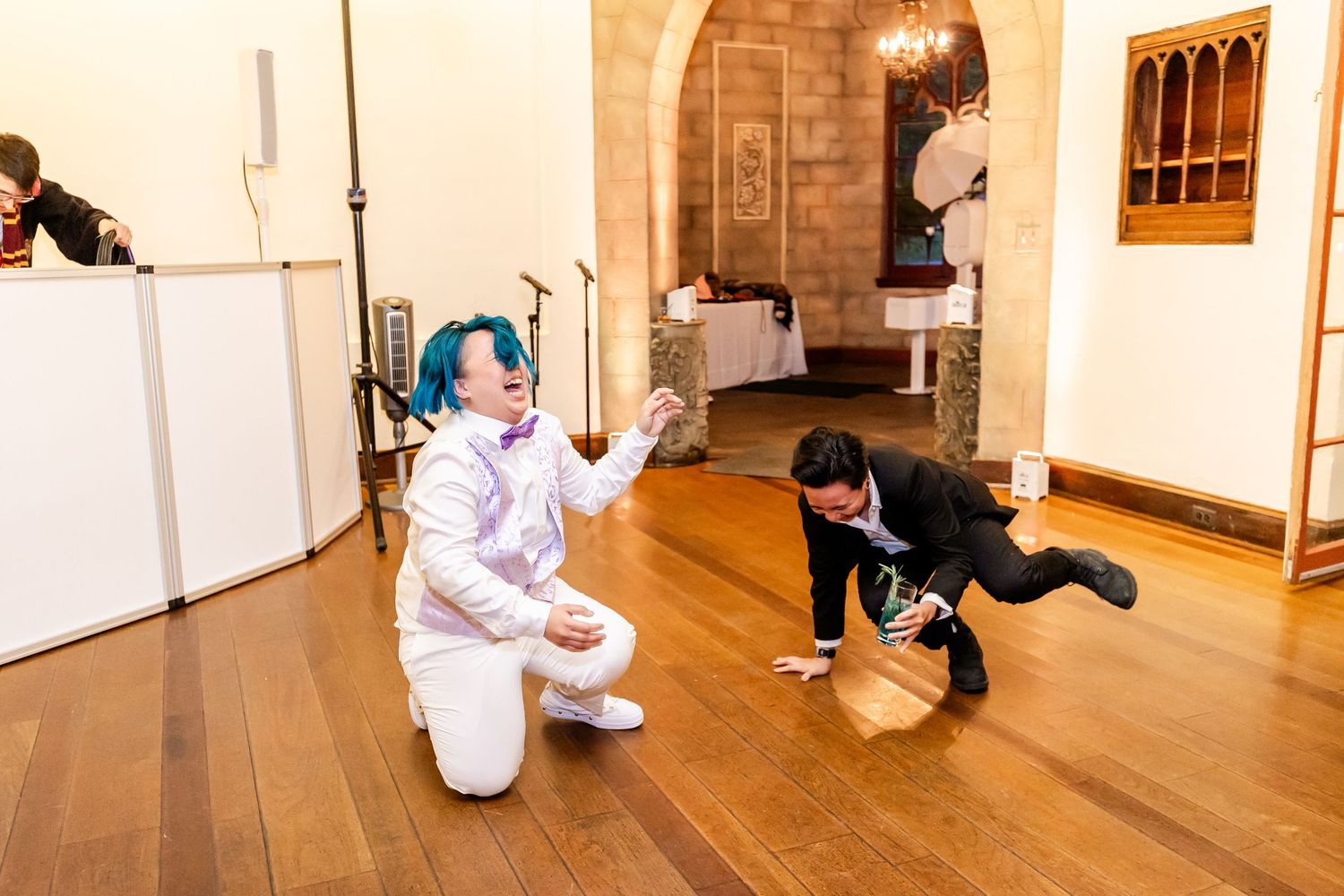 Performer in white outfit with blue hair dances while others watch at indoor celebration.