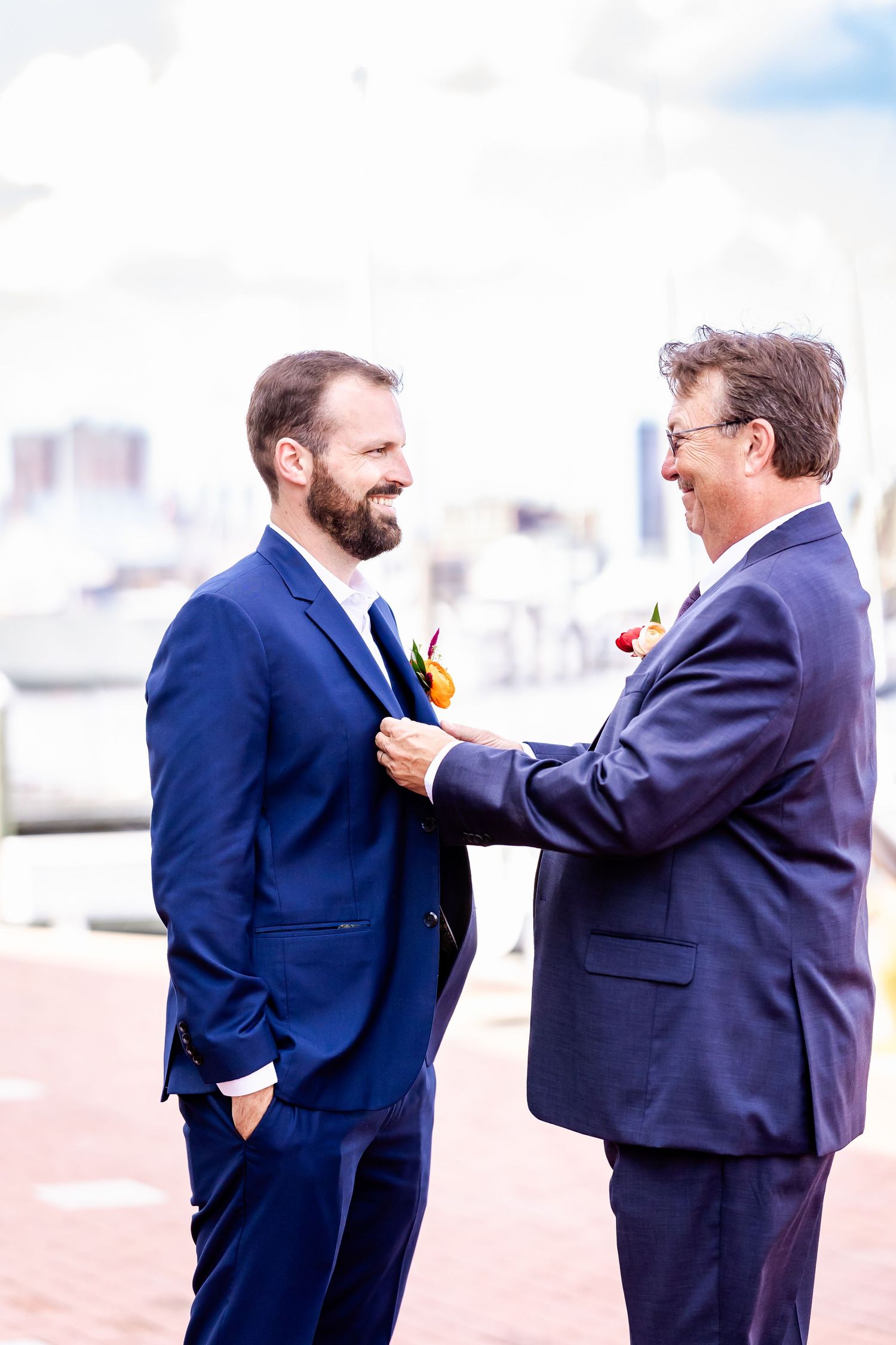 Two men in suits interact while standing outdoors with a city skyline in the background.