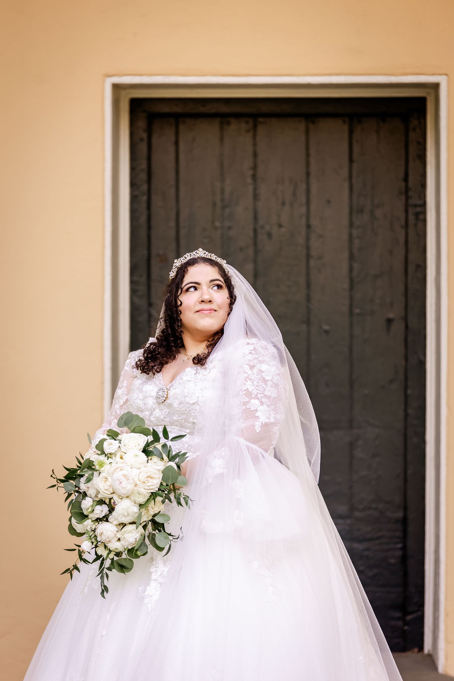 Bride with flowing veil holds white bouquet while posing against dark wooden doorway.