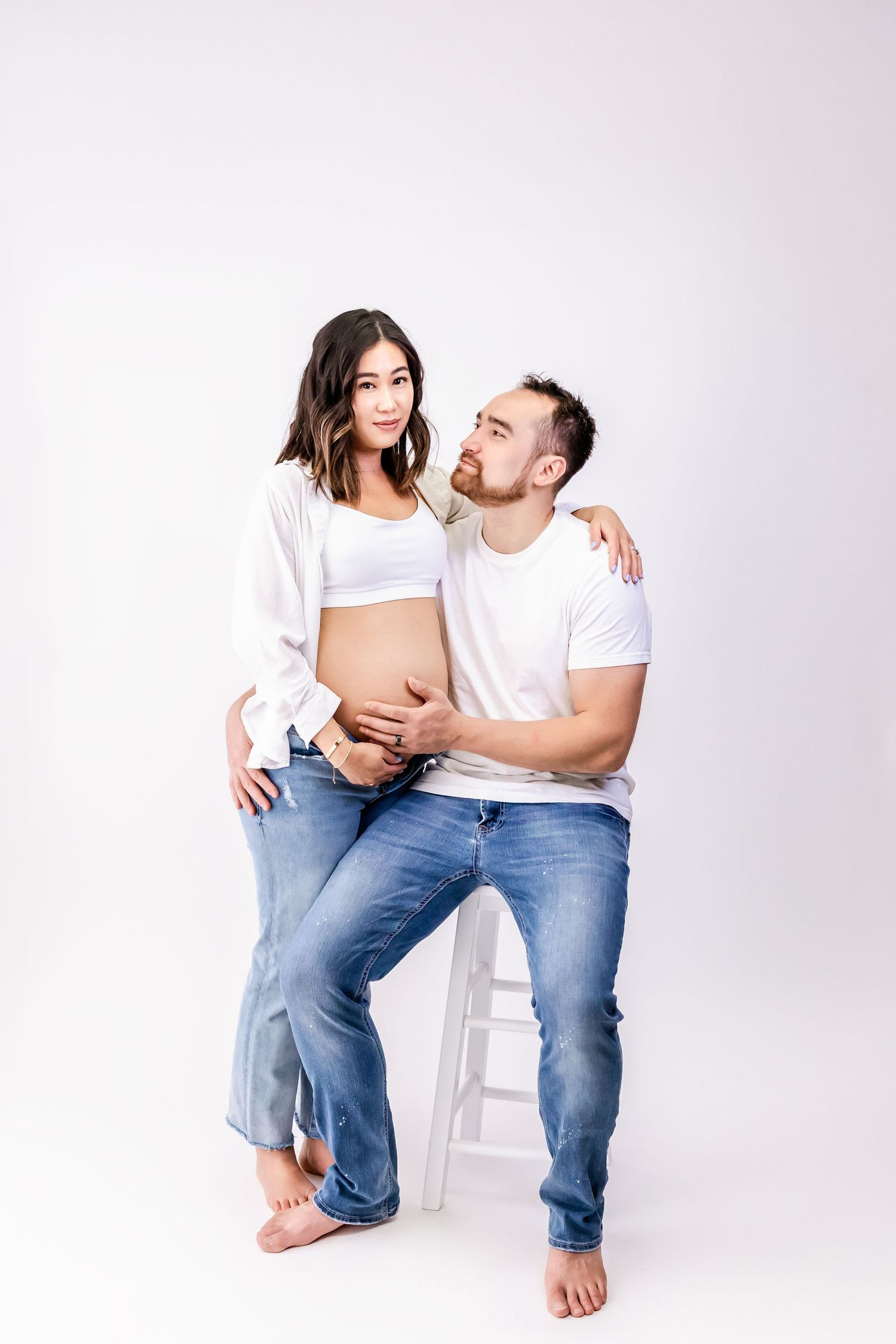 A couple in casual white tops and jeans pose together intimately against a light background during a maternity photoshoot.