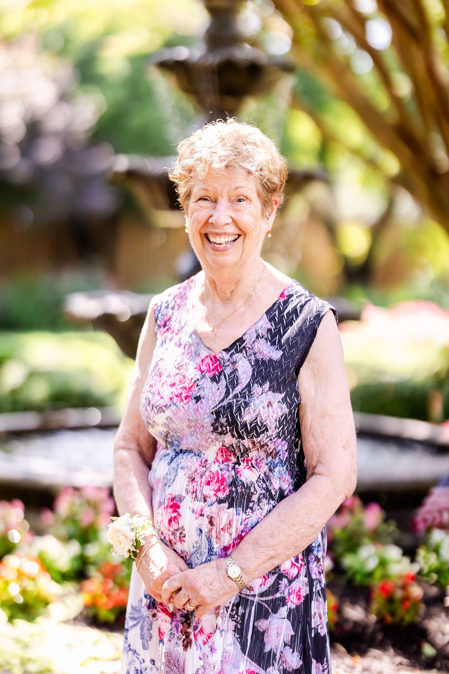 Woman in a floral dress smiling joyfully in a sunny garden with a fountain backdrop.