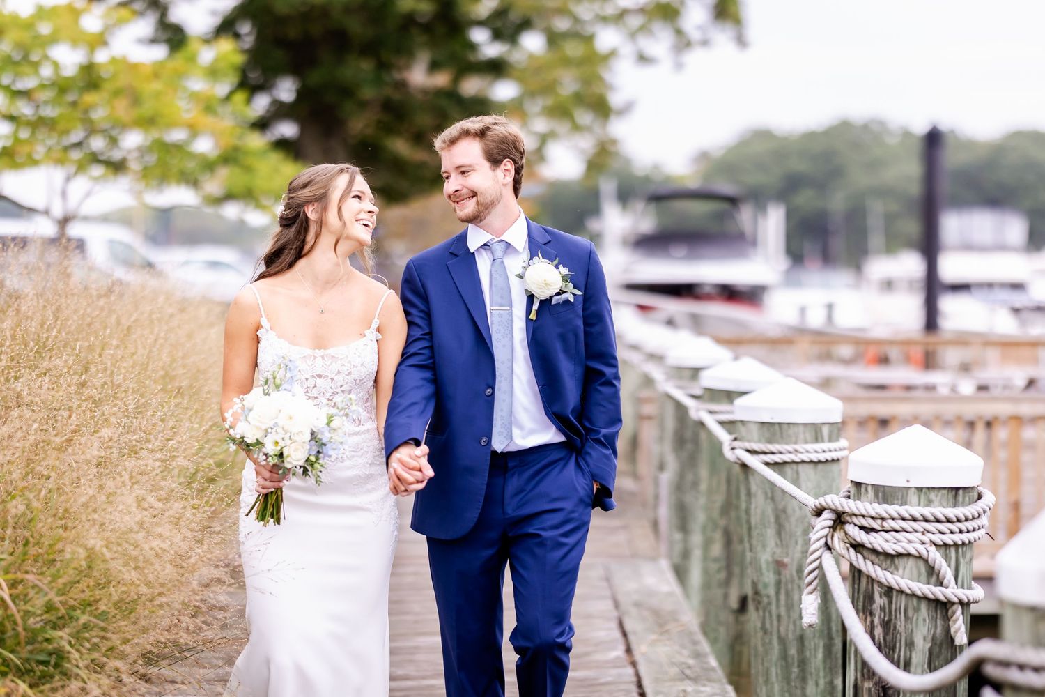 A couple in wedding attire walks along a marina dock with boats and tall grass in the background.
