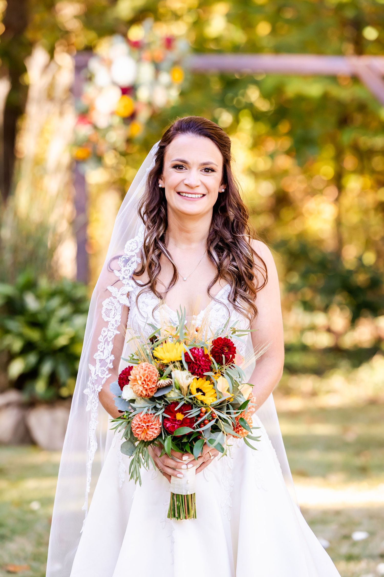 Elegant bride in white lace gown holding vibrant fall-colored bouquet with roses and greenery.