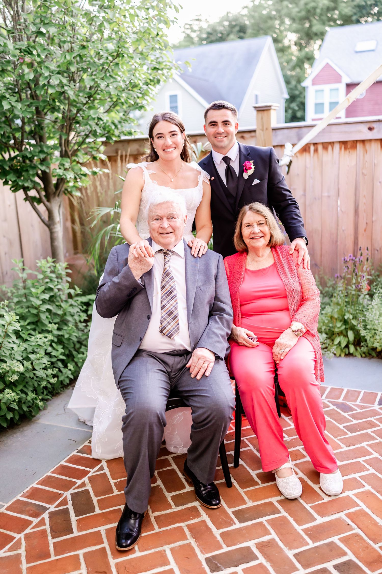 Couple sits together on brick steps in a garden setting with greenery and wooden fence in background.