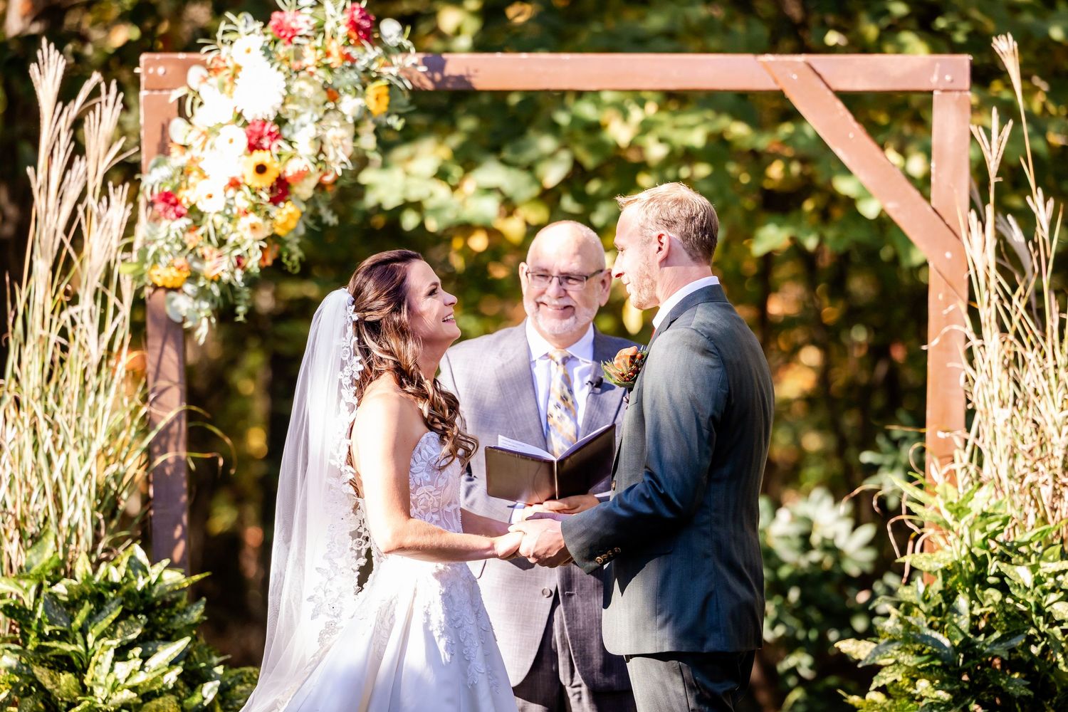 Wedding ceremony takes place in a beautiful outdoor garden setting with autumn foliage and decorative arch.